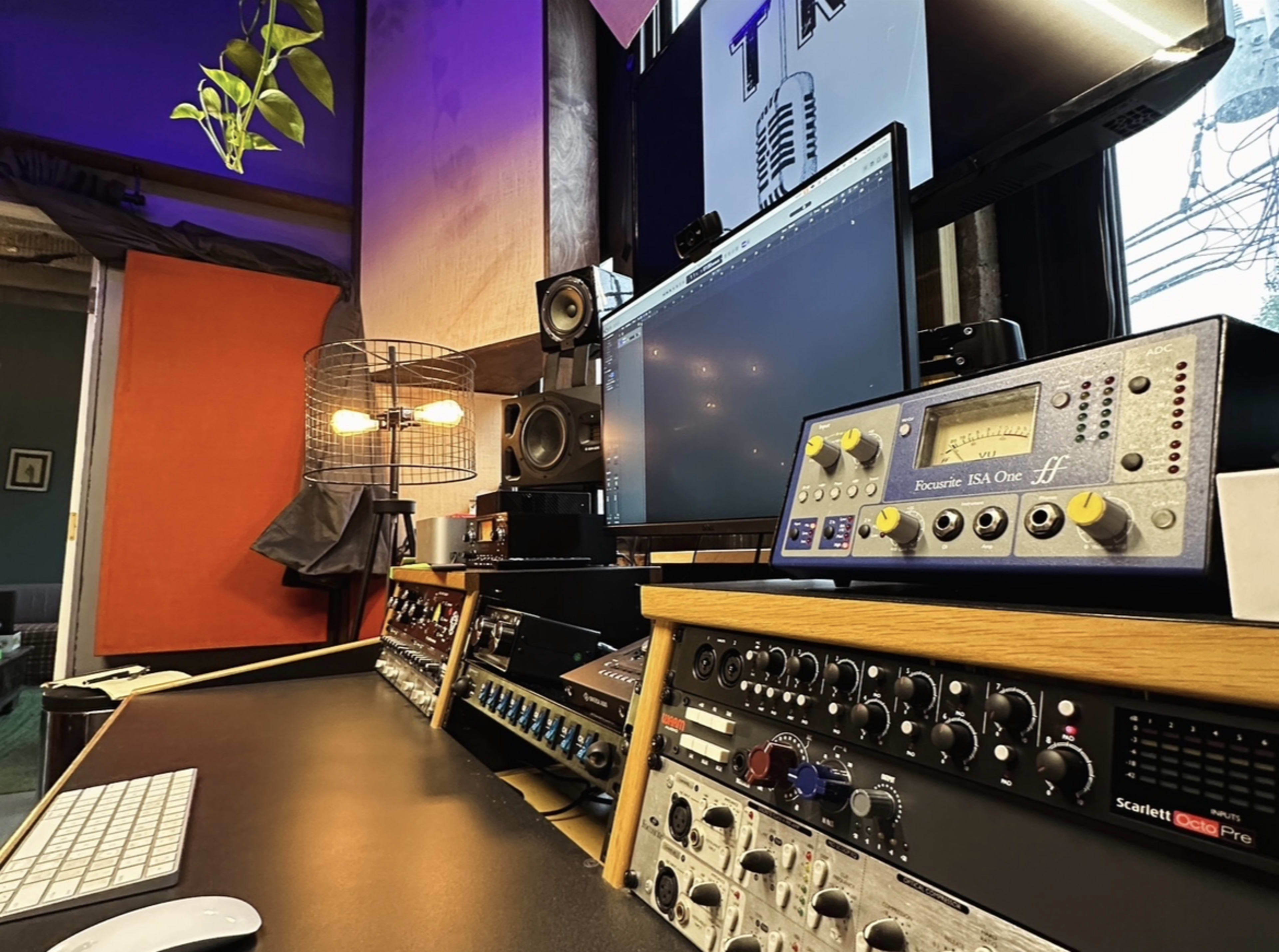 The image shows a recording studio workspace with a computer monitor, audio equipment, and a keyboard on a desk, alongside various sound mixers and an overhead plant.