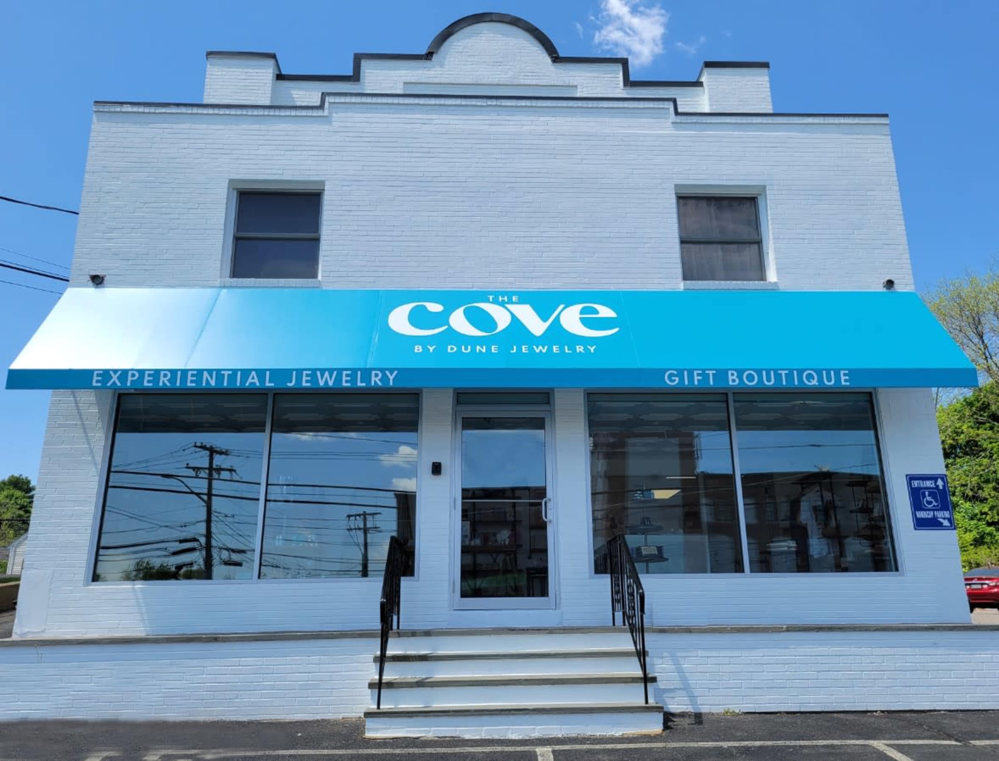 A white brick building with a large blue awning displaying the name "The Cove by Dune Jewelry," which serves as a gift boutique.