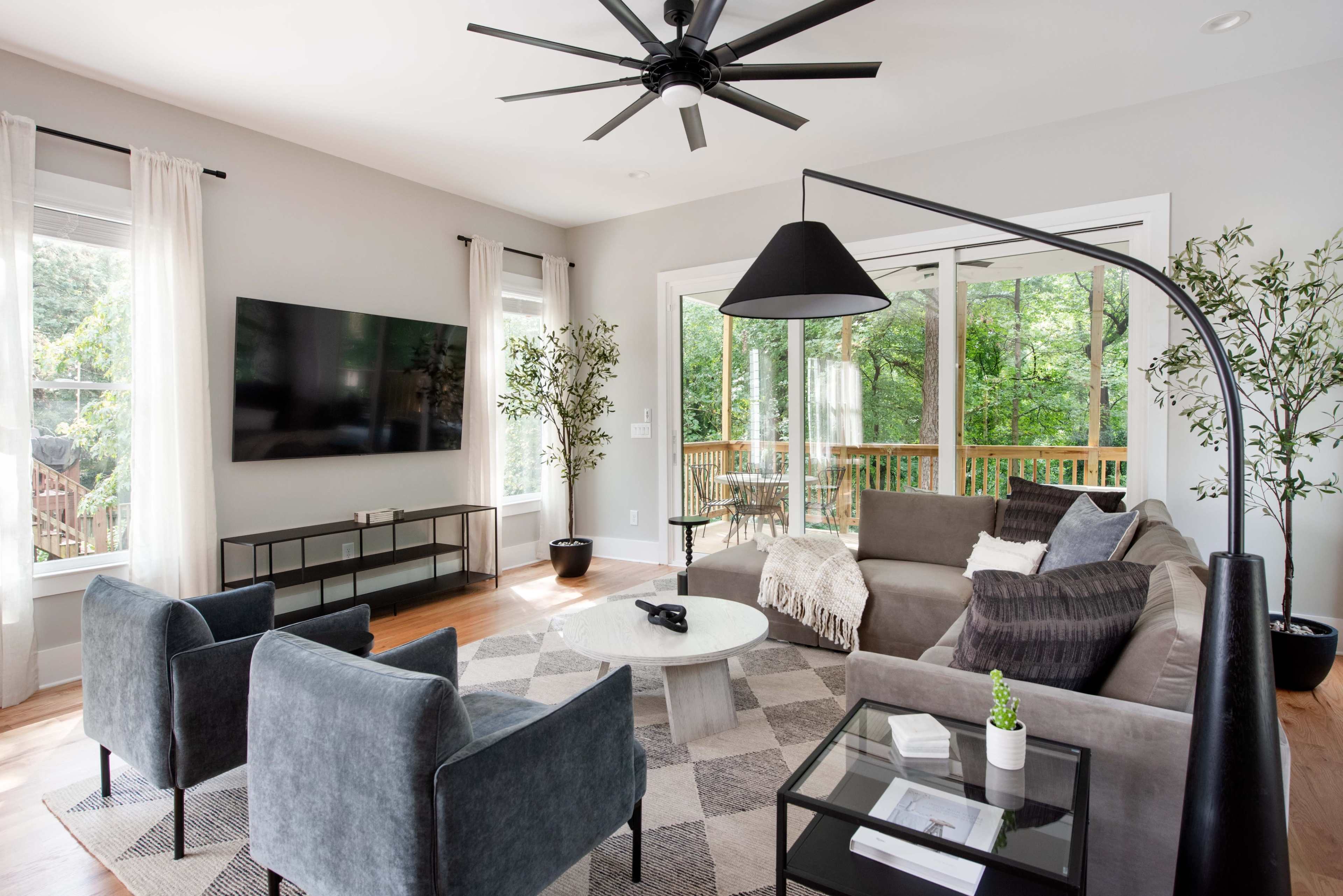A contemporary living room features a sectional sofa, two armchairs, a coffee table, and a large television, all set against large windows that overlook greenery.