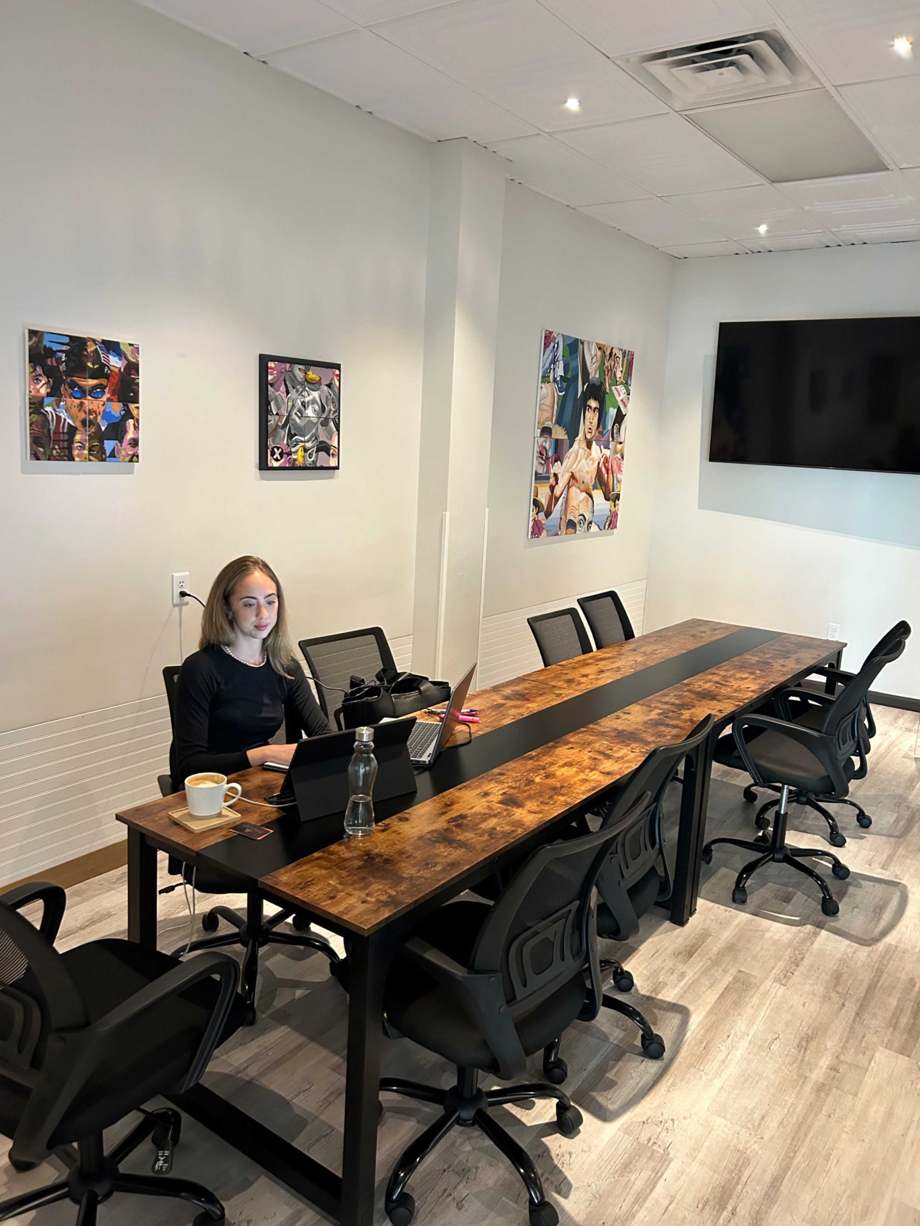 A woman sits at a large, modern conference table with a laptop and a cup, while colorful artwork adorns the walls of the bright meeting room.
