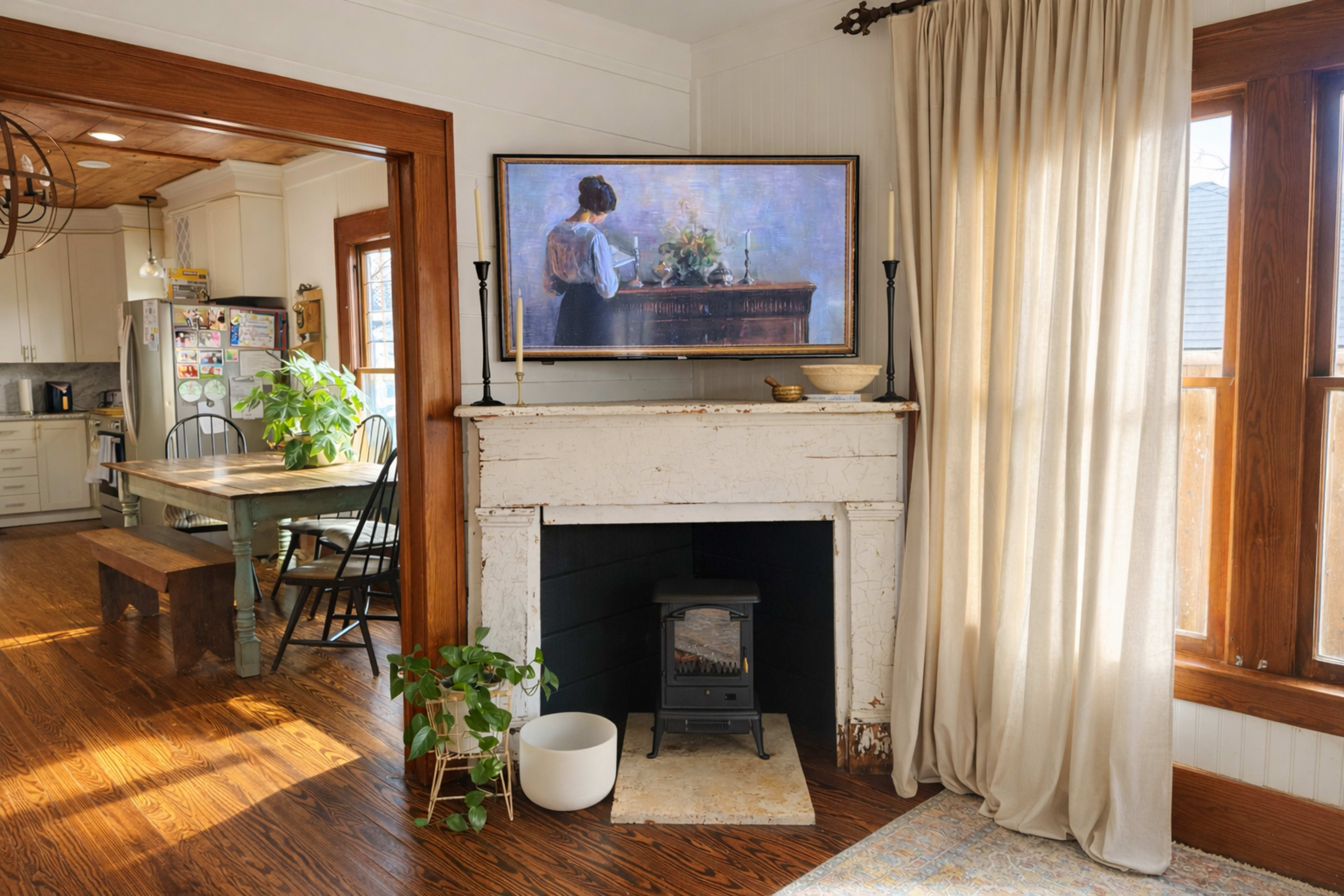 The image shows a cozy interior space featuring a rustic fireplace with a small stove and a painting above it, alongside a dining area with a wooden table and chairs.