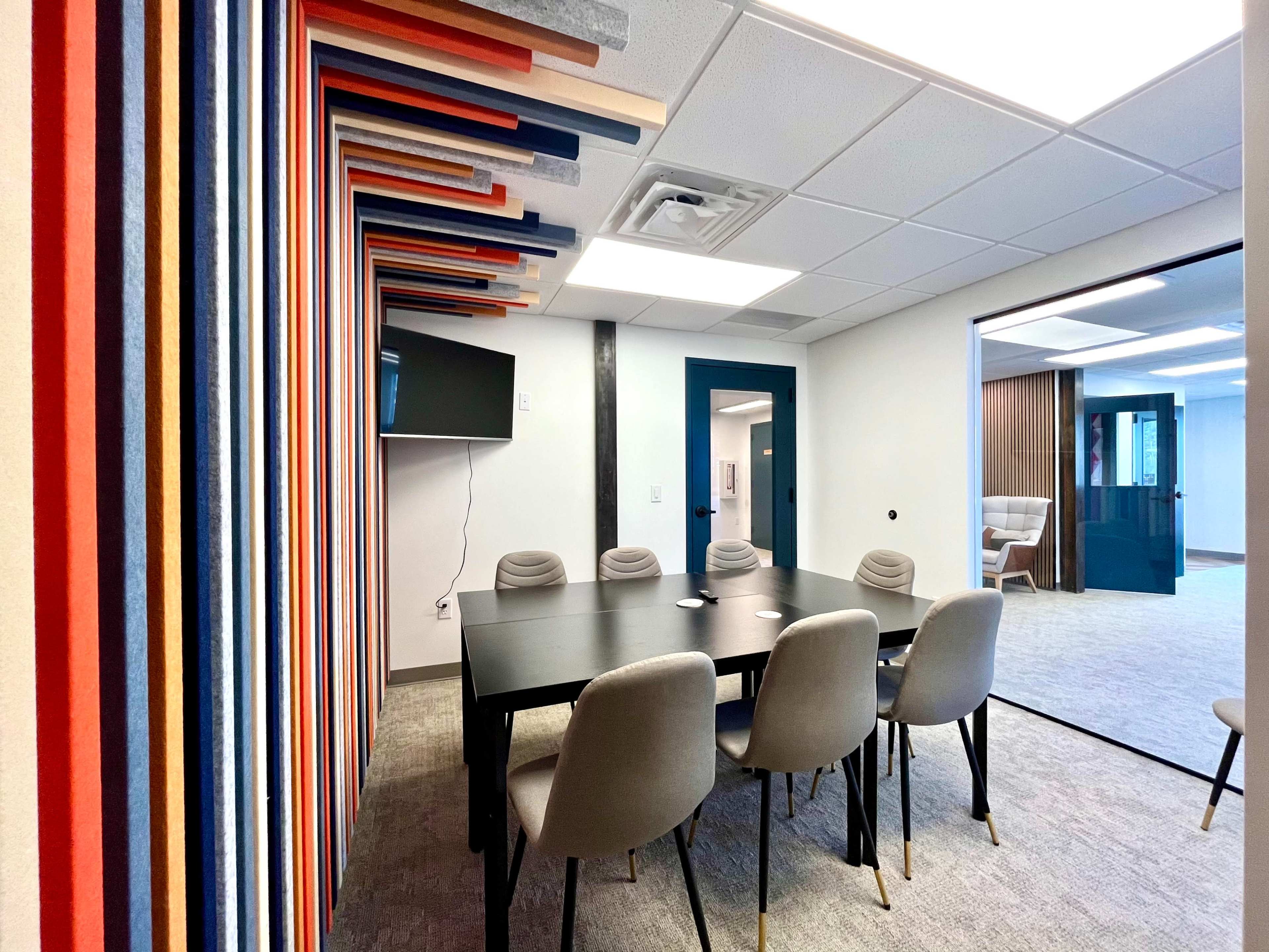 A modern conference room featuring a black table surrounded by gray chairs, with a colorful striped wall and a wall-mounted television.