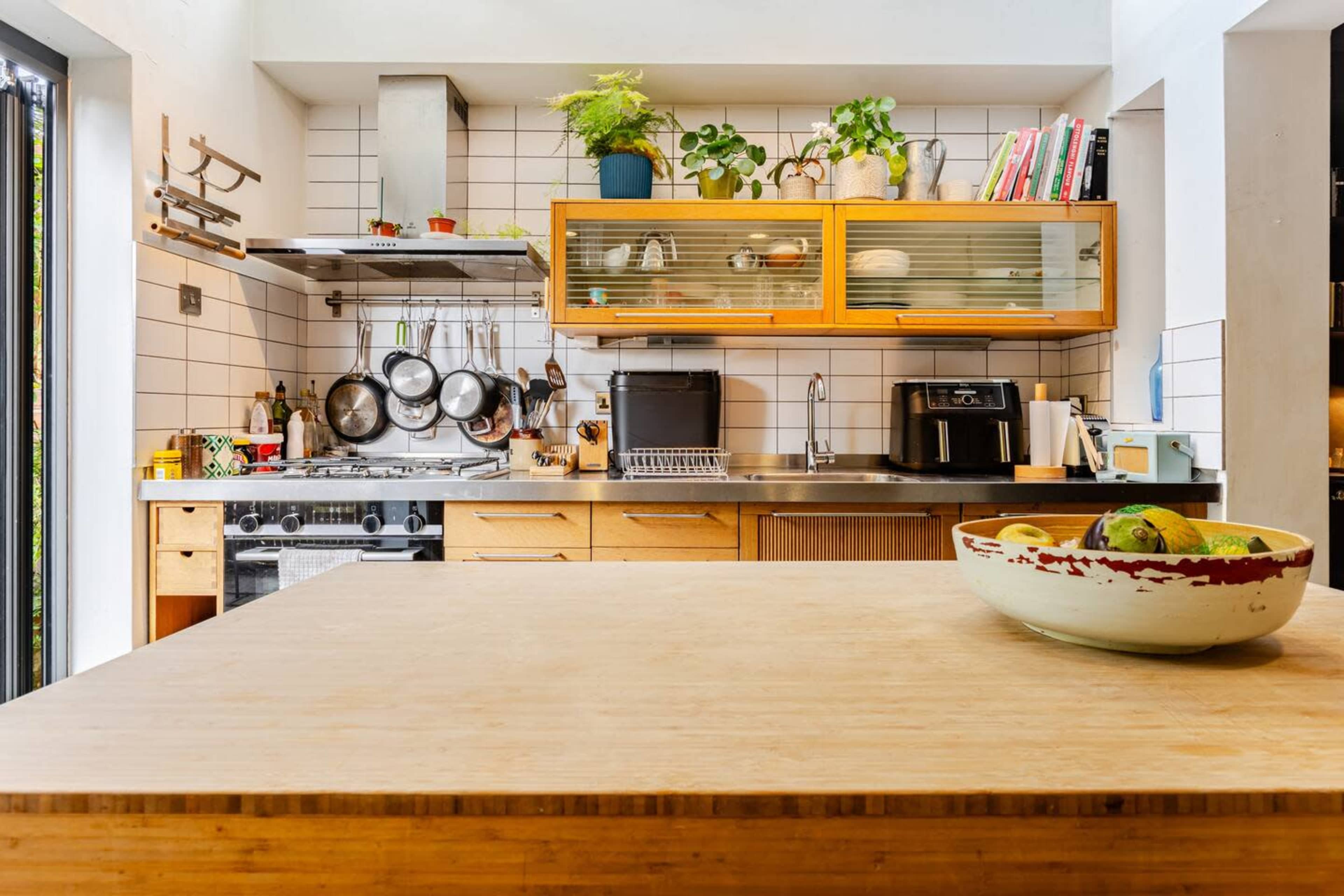 A modern kitchen features a wooden island with a bowl of fruit, a stove, hanging pots, and shelves with plants and cookbooks.