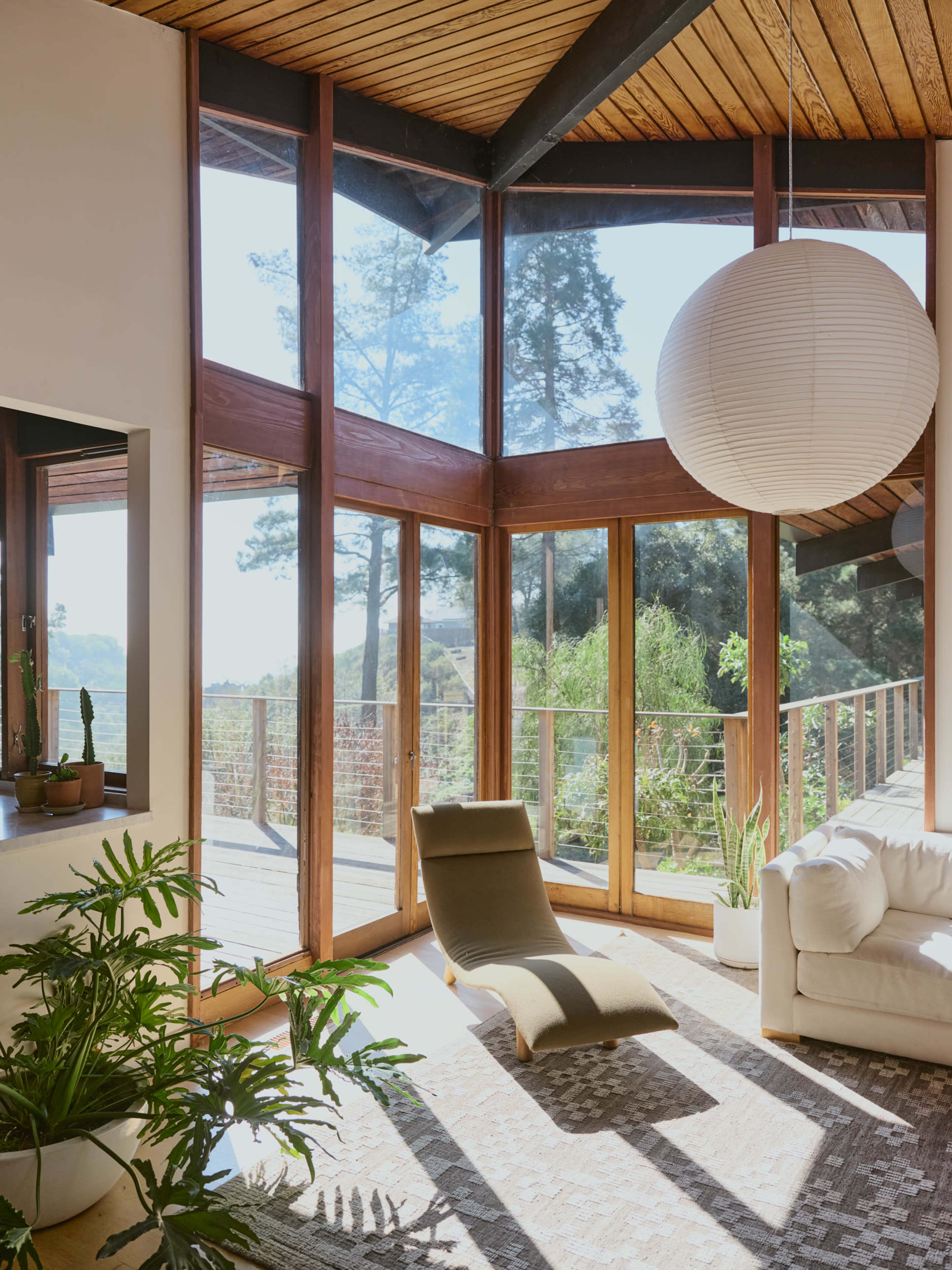 A sunlit living room features large windows, a modern lounge chair, a white sofa, and indoor plants, creating a connection to the outdoors.