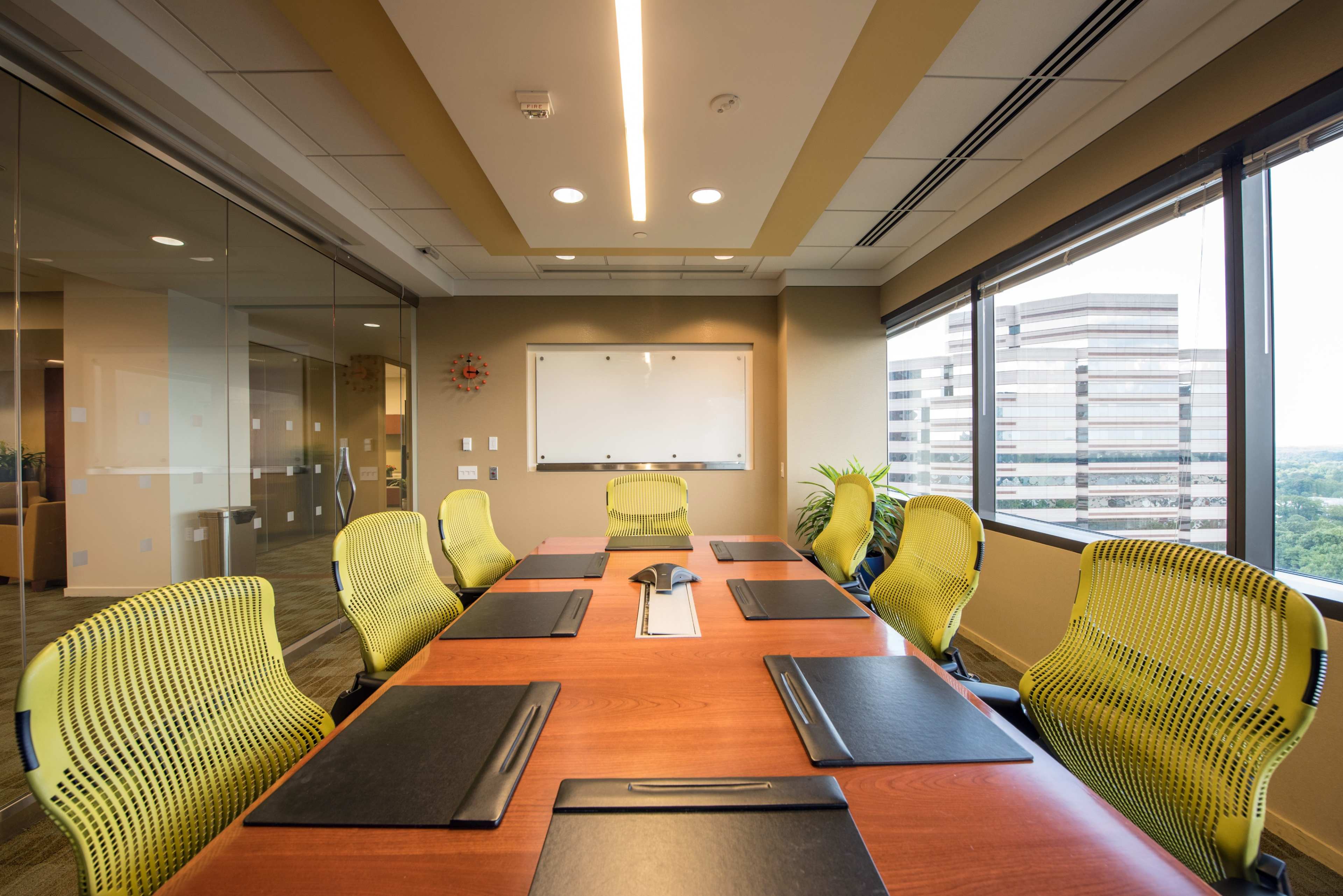 A modern conference room features a large wooden table surrounded by green ergonomic chairs, with a whiteboard and windows overlooking a cityscape.