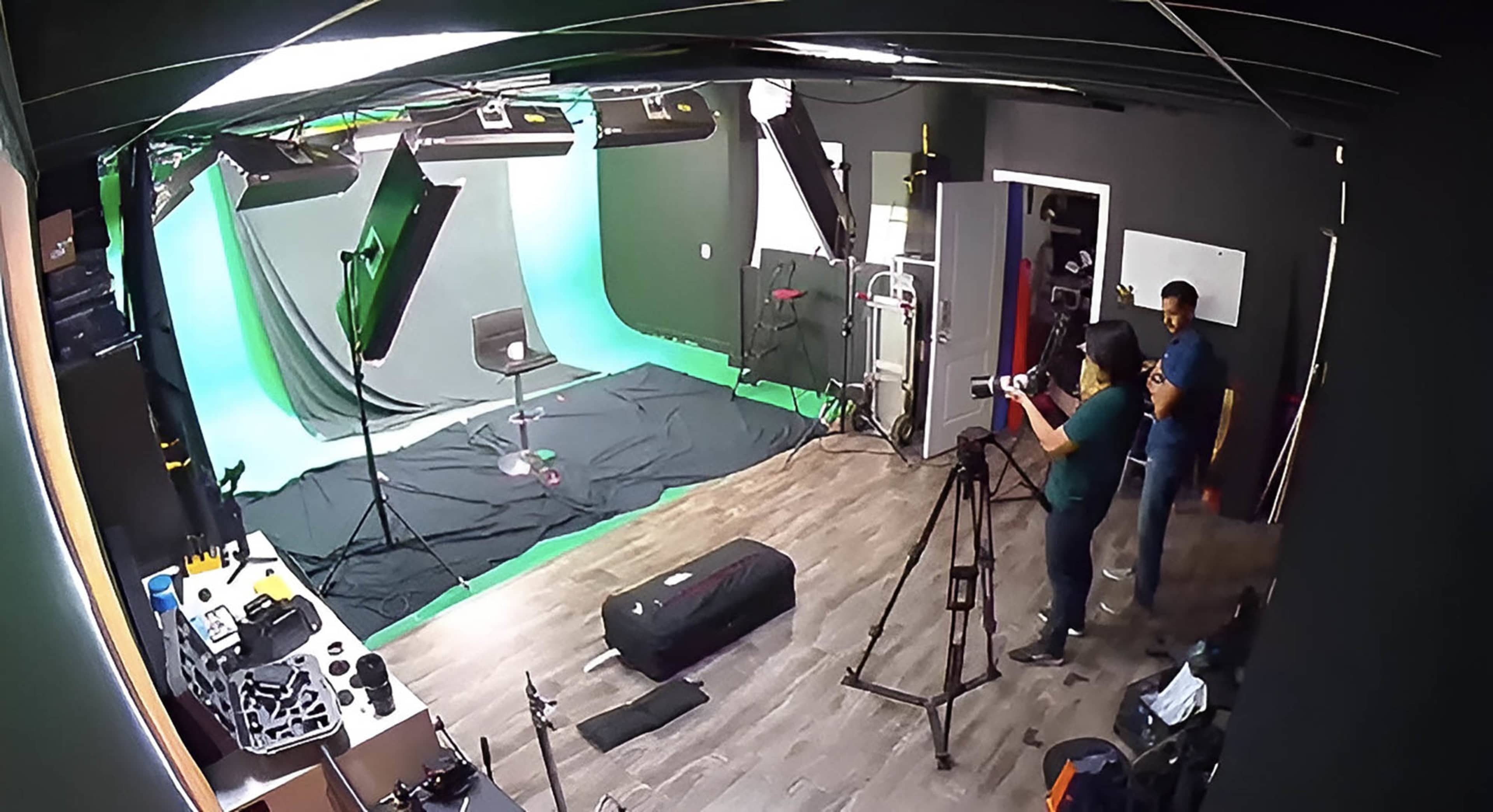 A video production studio is set up with a green screen, lighting equipment, and a camera on a tripod, while two individuals prepare for a shoot.