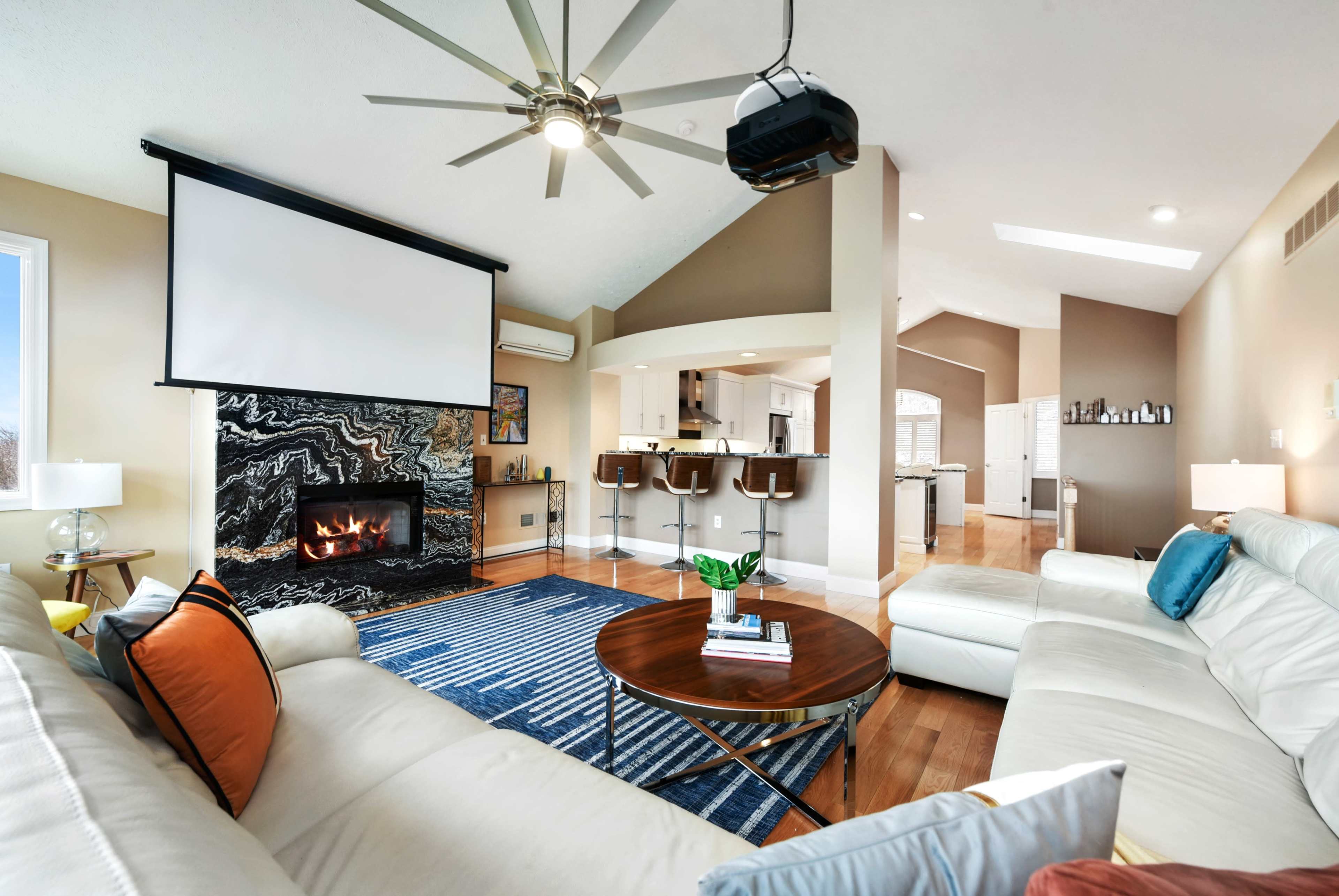 The living room features a white sofa set, a large projection screen, a stone fireplace, and a coffee table, all arranged around a patterned area rug.