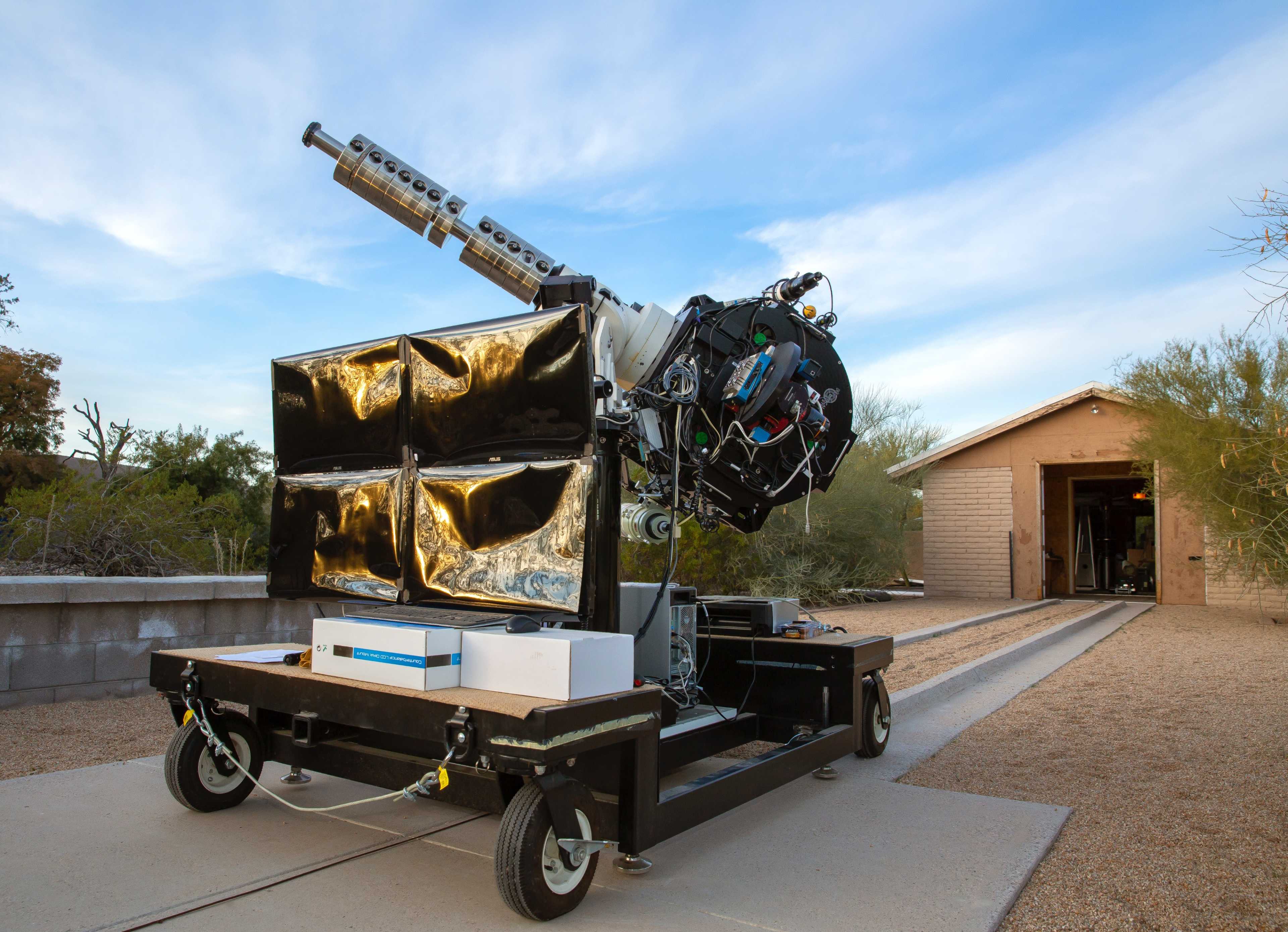 A scientific instrument on a wheeled platform is positioned outside a building, with a long arm extending upwards toward the sky.