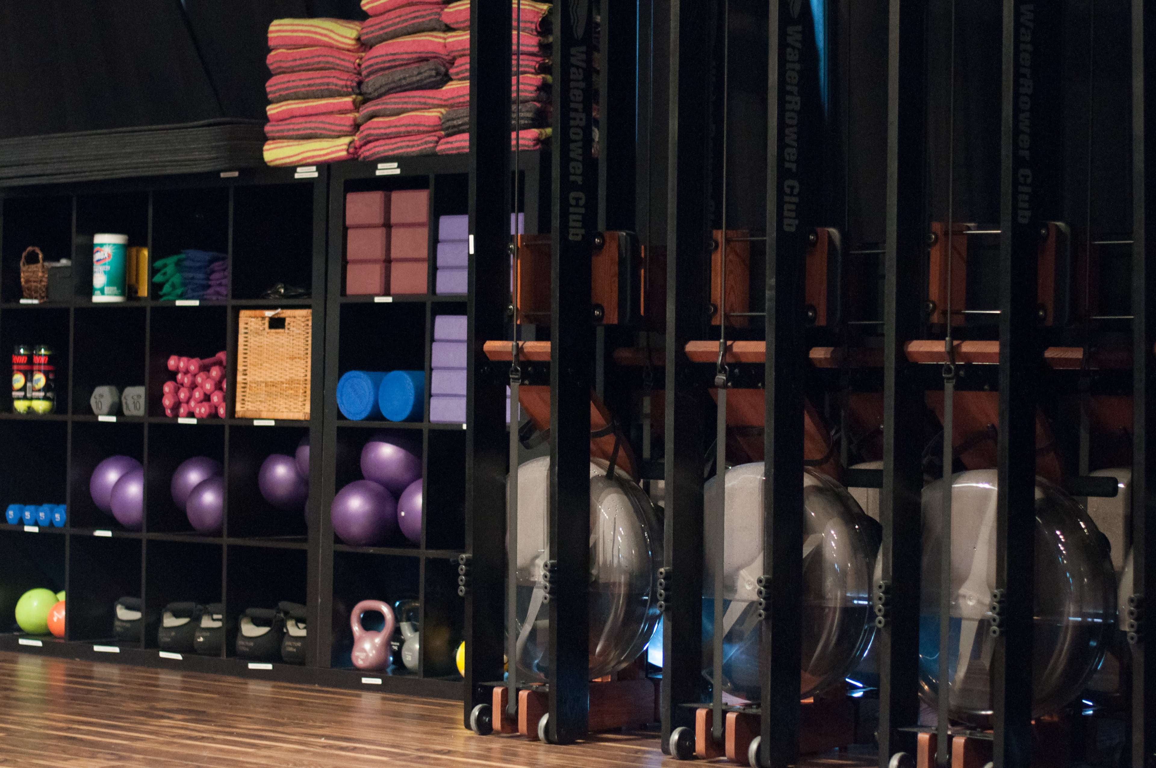 The image shows a fitness studio with organized shelves containing exercise equipment, including weights and resistance bands, alongside neatly stacked towels.