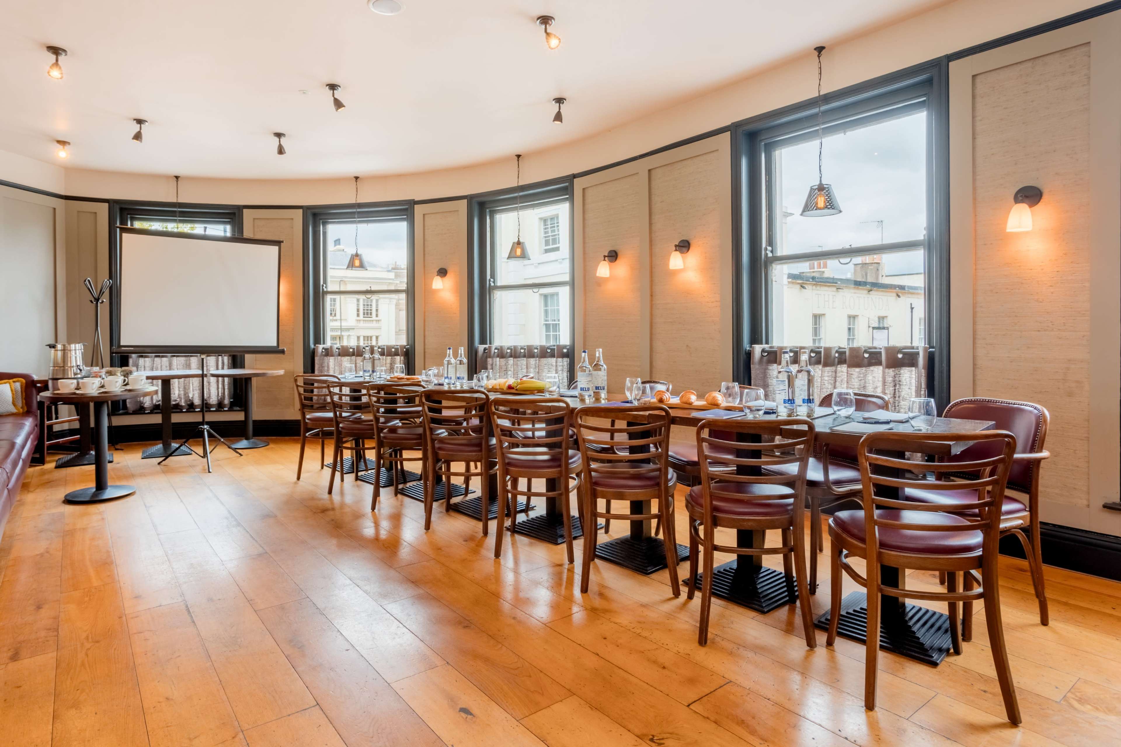 A conference room with a long rectangular table set for a meeting, featuring large windows and simple wooden chairs.