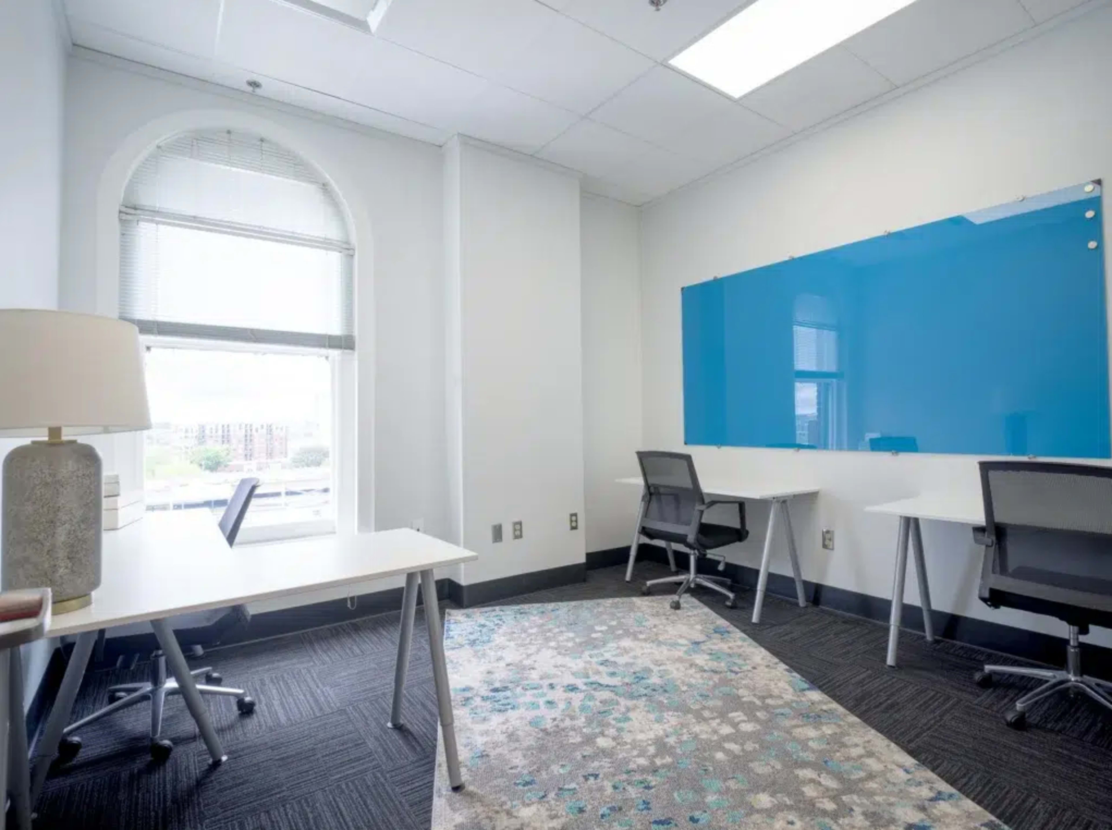 The image shows a bright office space with two desks facing a large window and a blue whiteboard on the wall.