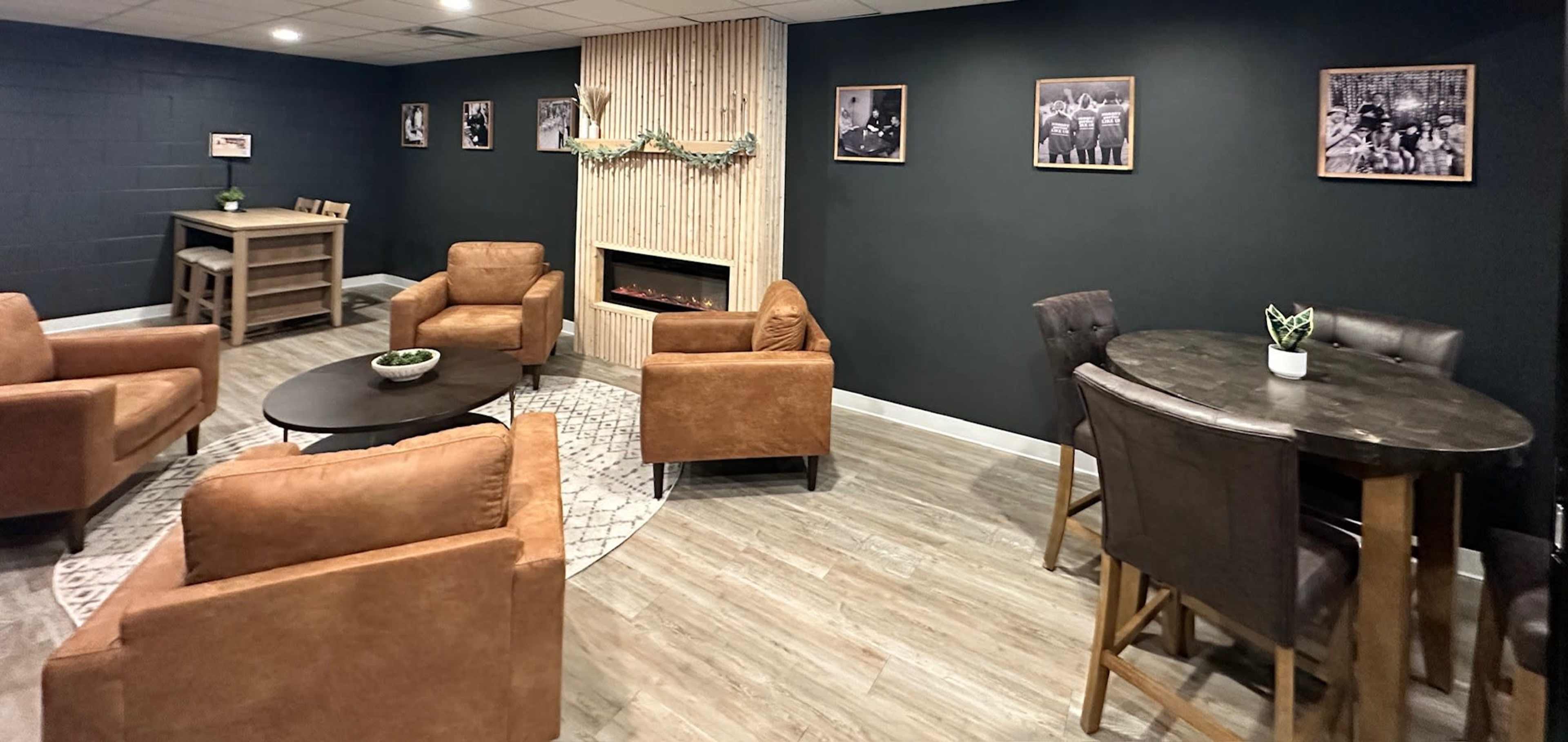A cozy lounge area featuring leather armchairs, a round table with upholstered chairs, a wall-mounted electric fireplace, and framed photographs on the walls.