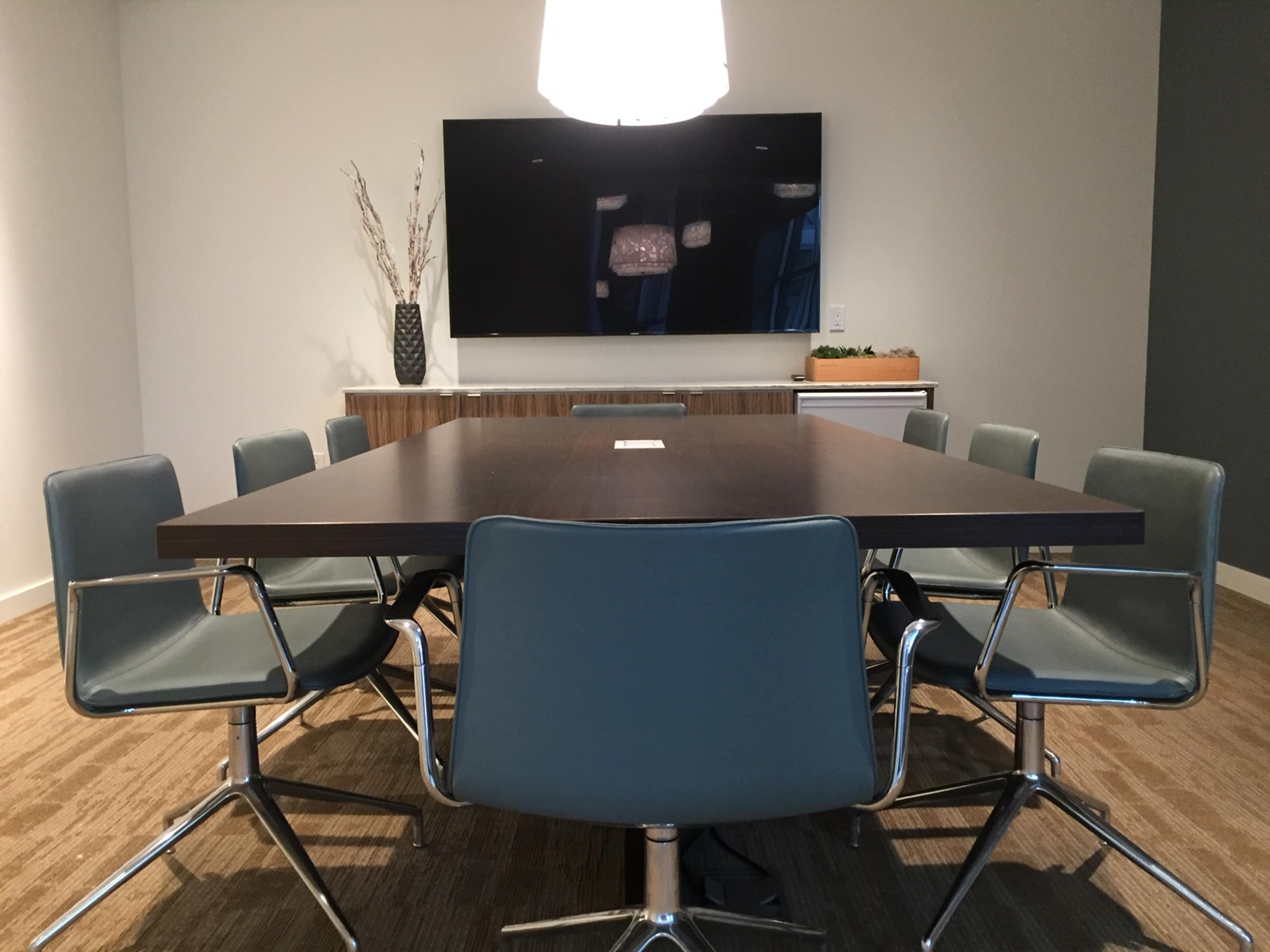 A modern conference room features a large rectangular table surrounded by several chairs and a wall-mounted screen.