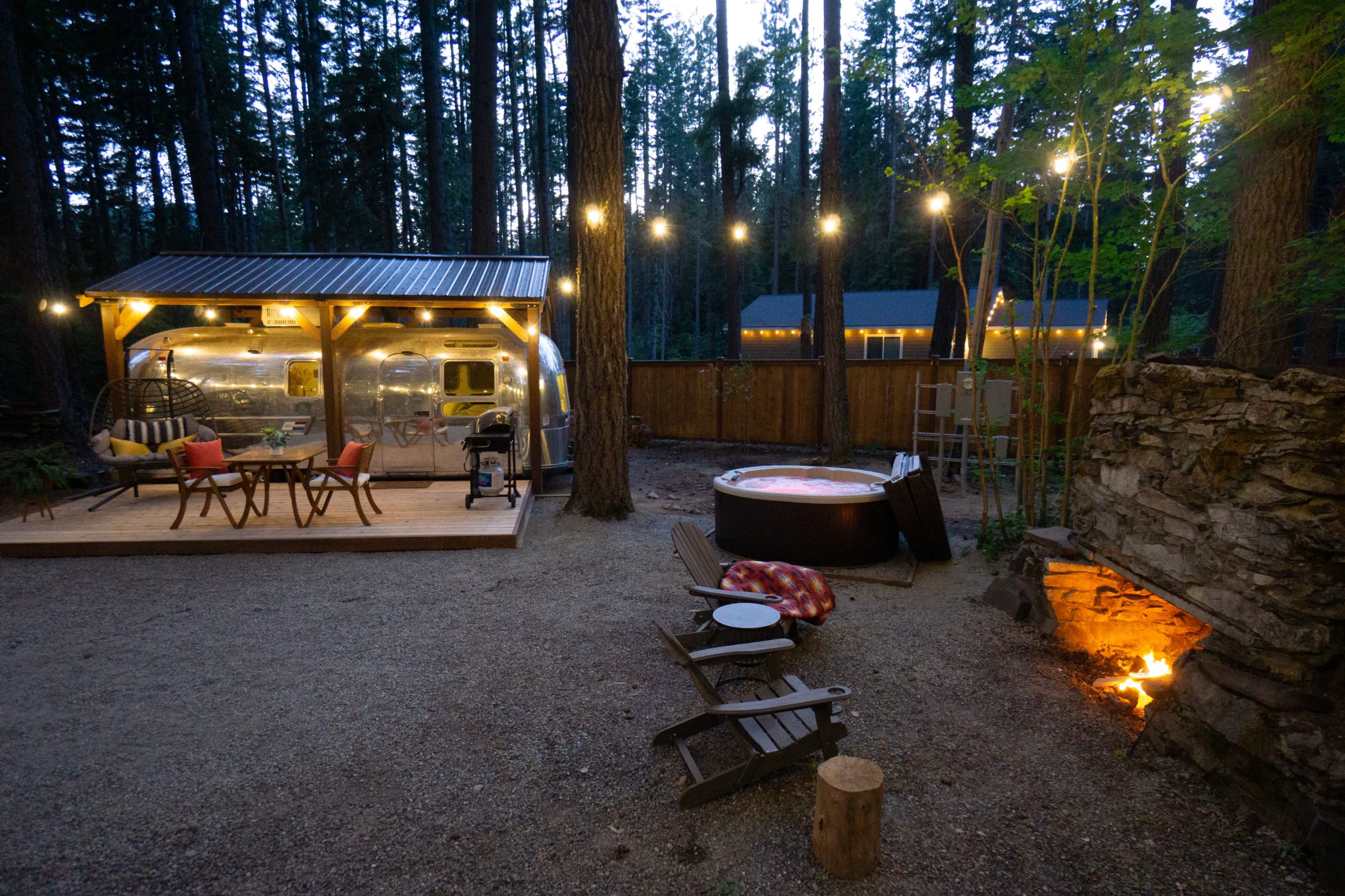 A cozy outdoor seating area with string lights, a hot tub, and a campfire is set up beside a weathered trailer in a forested campsite.