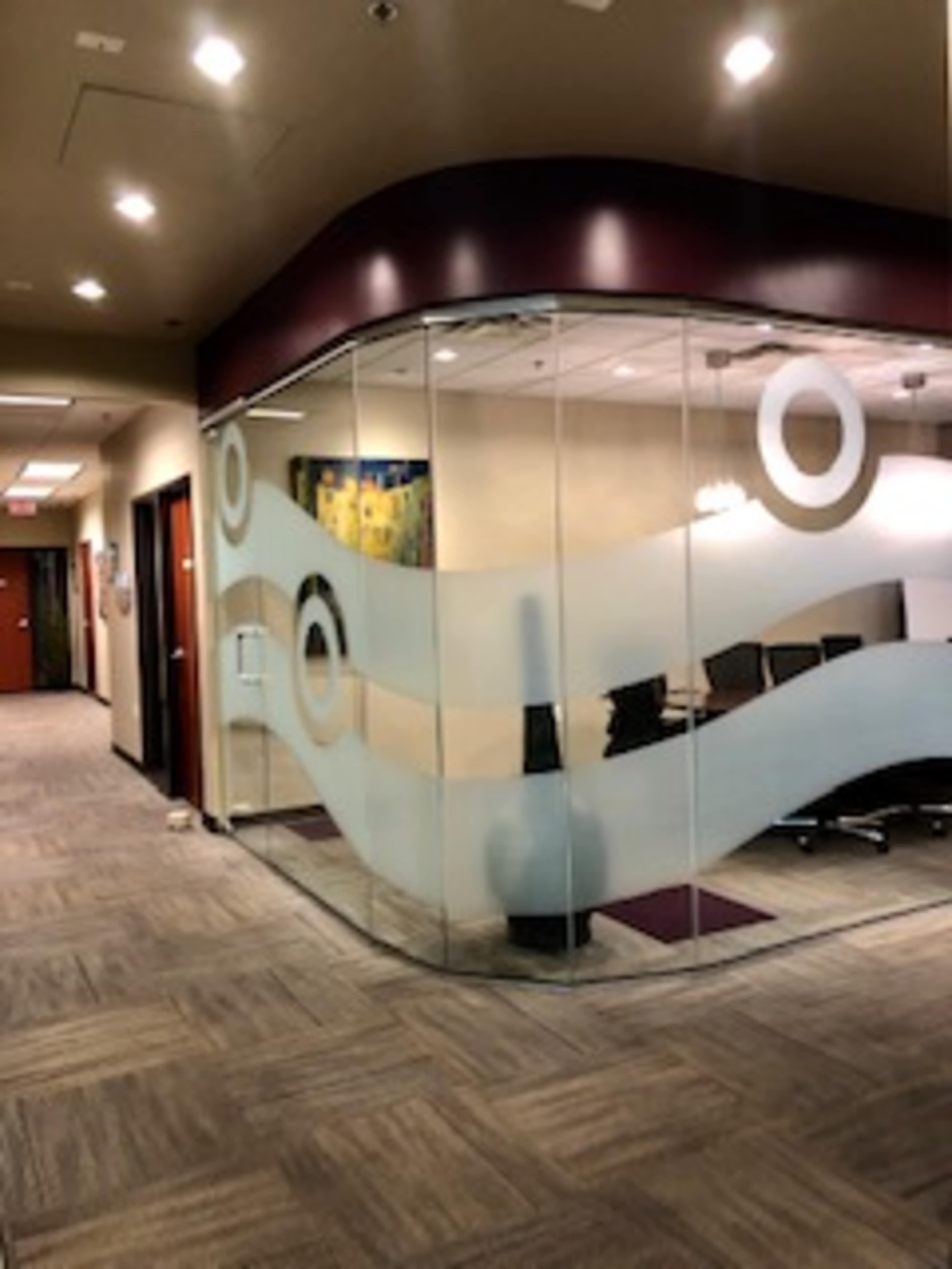 The image shows a modern office space with a glass-walled conference room featuring curved frosted designs and a carpeted entrance.