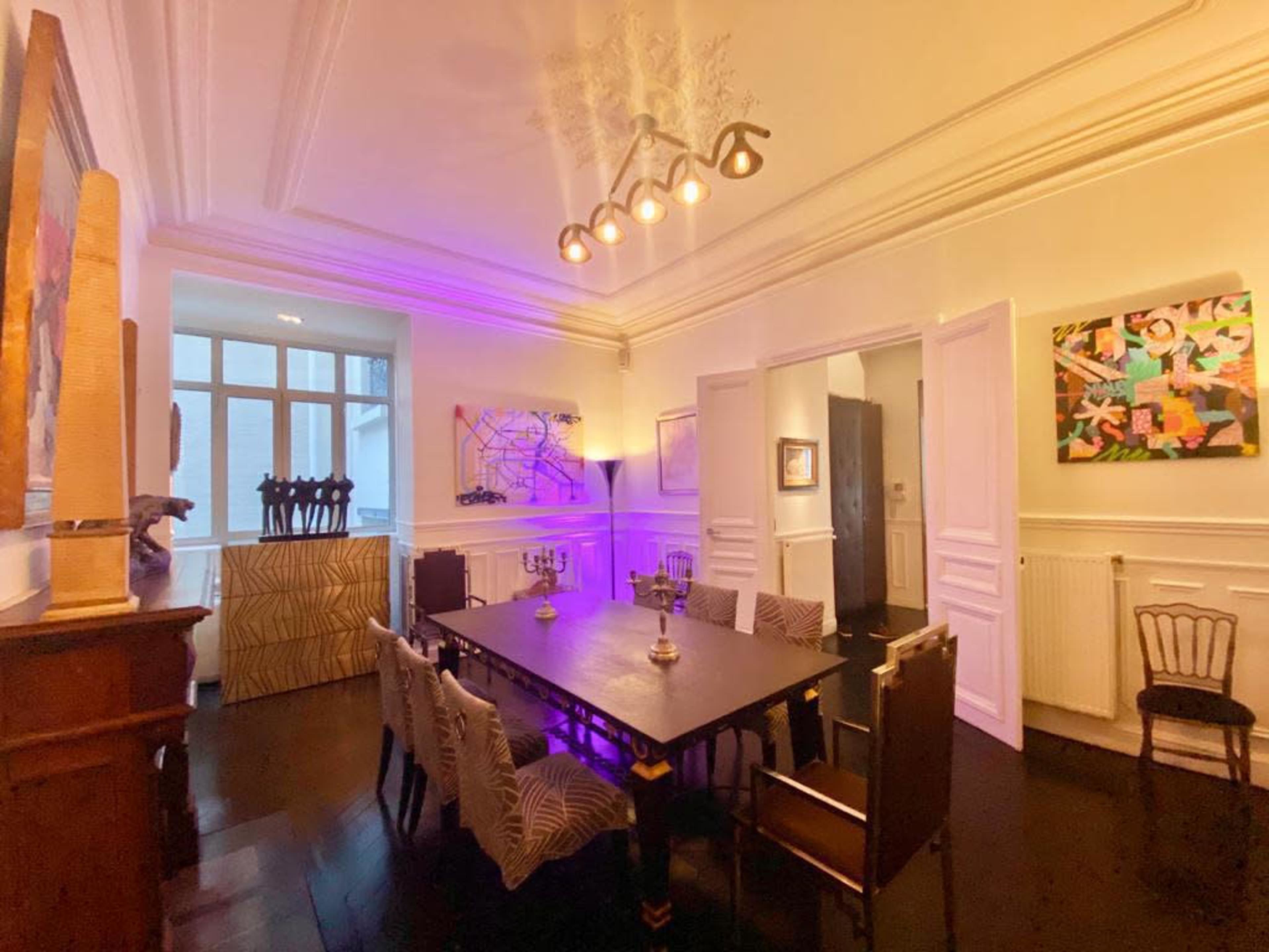 A dining room features a large table surrounded by chairs, with artwork on the walls and modern lighting overhead.