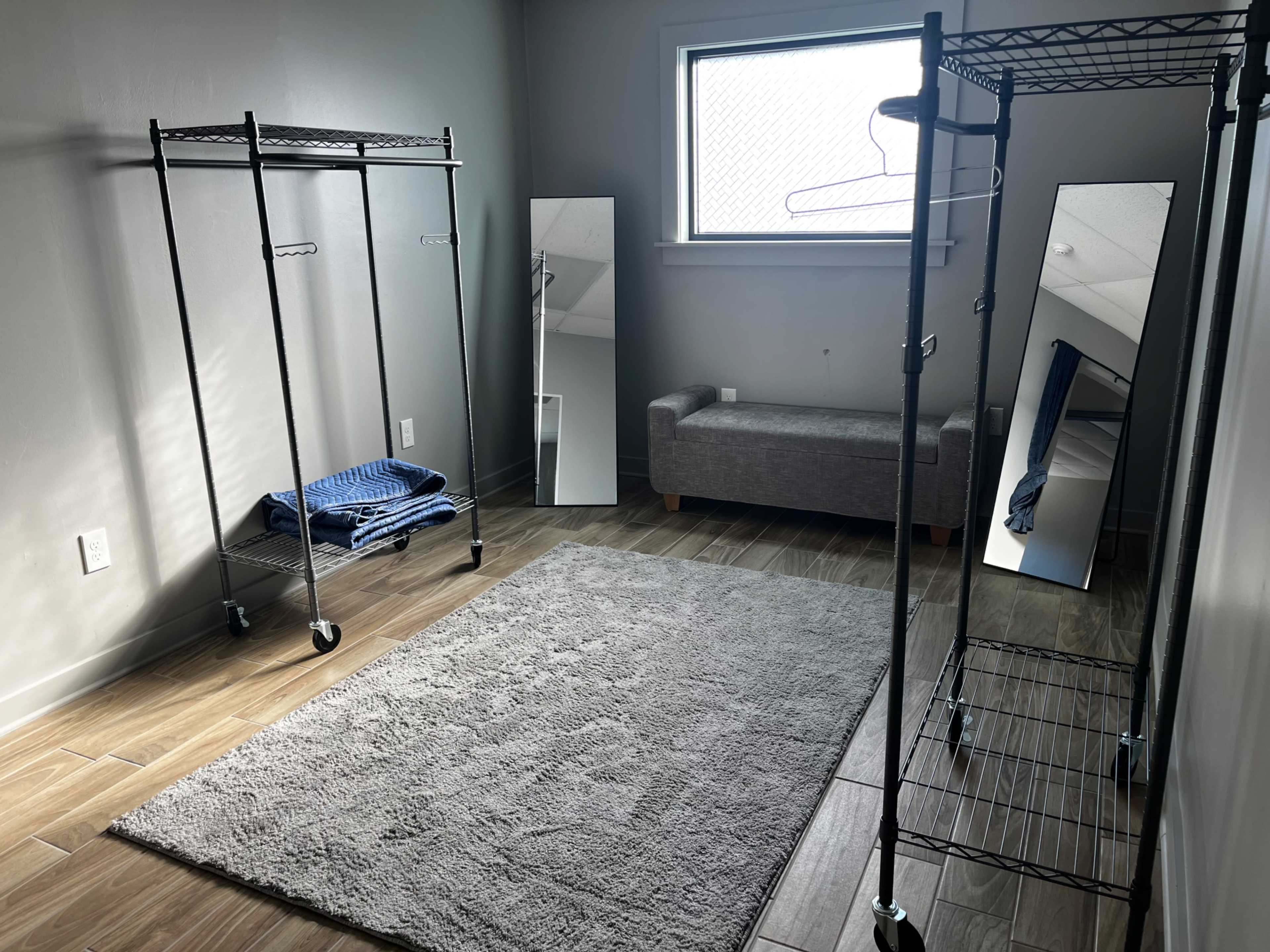 The image shows a minimalist room with two clothing racks, a gray sofa, two mirrors, and a light gray rug on a wood-like floor.