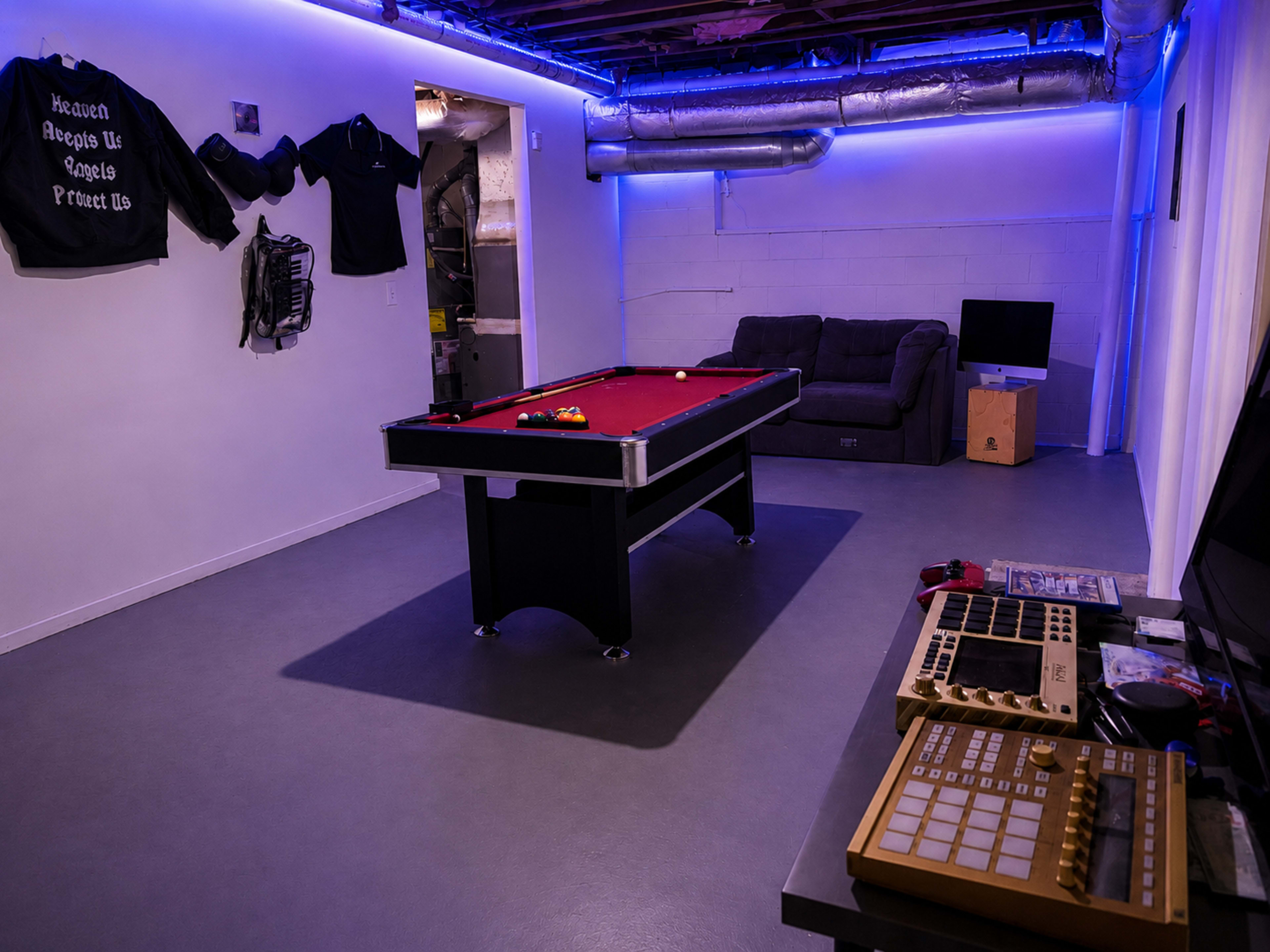 The image depicts a basement recreation room featuring a pool table, a couch, a TV, and various items on a workspace.
