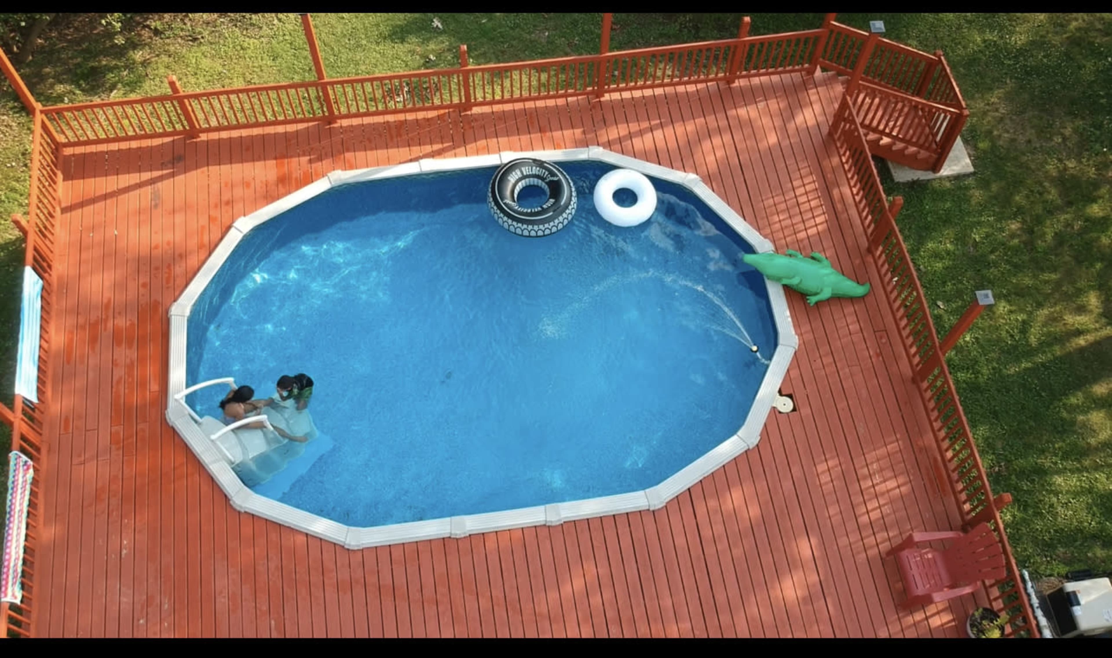 The image shows an aerial view of a backyard pool surrounded by a wooden deck, with two people in the water and floating toys nearby.