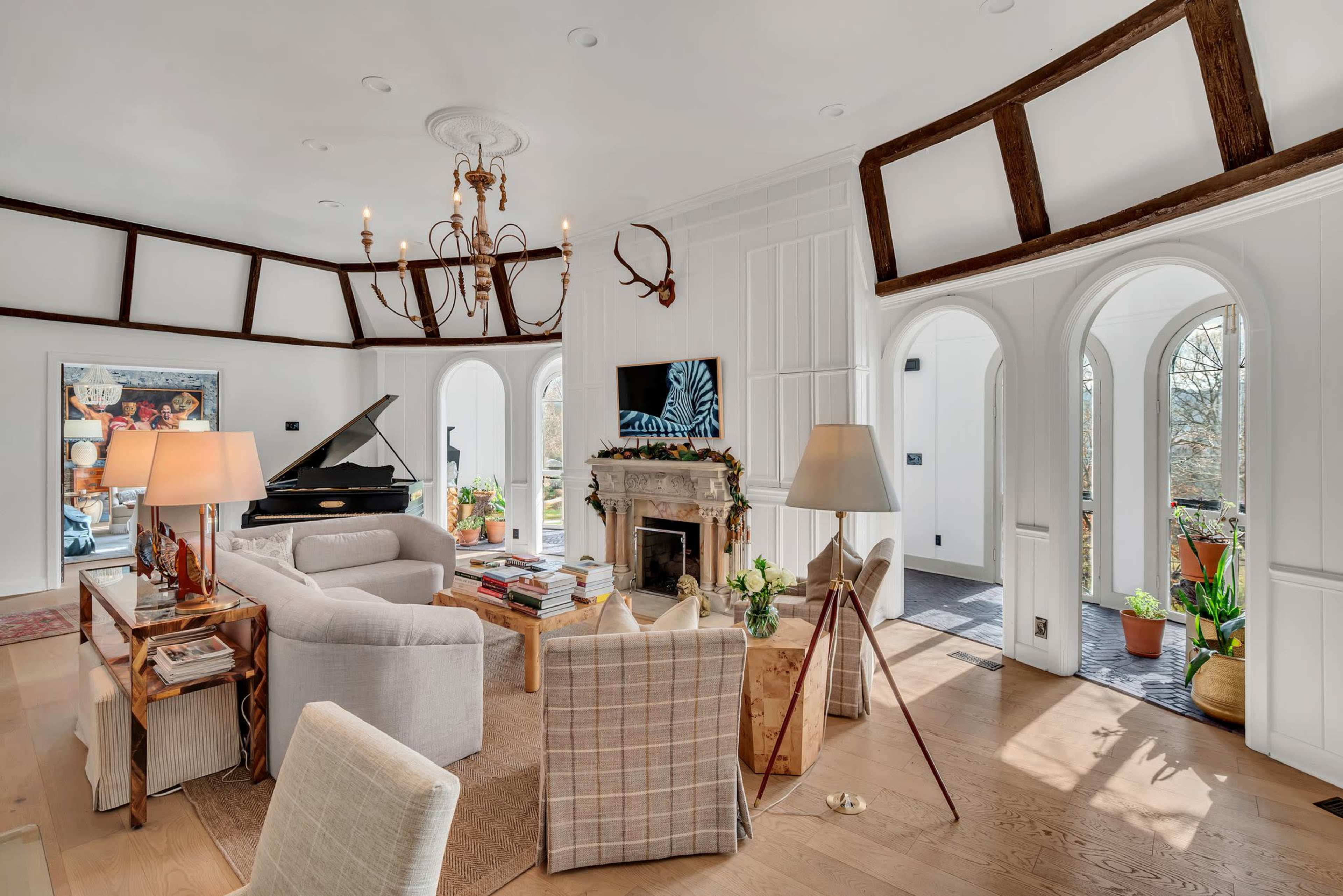 The image shows a modern living room with a grand piano, a fireplace, and large windows that provide natural light, featuring a mix of furniture arranged around a central space.