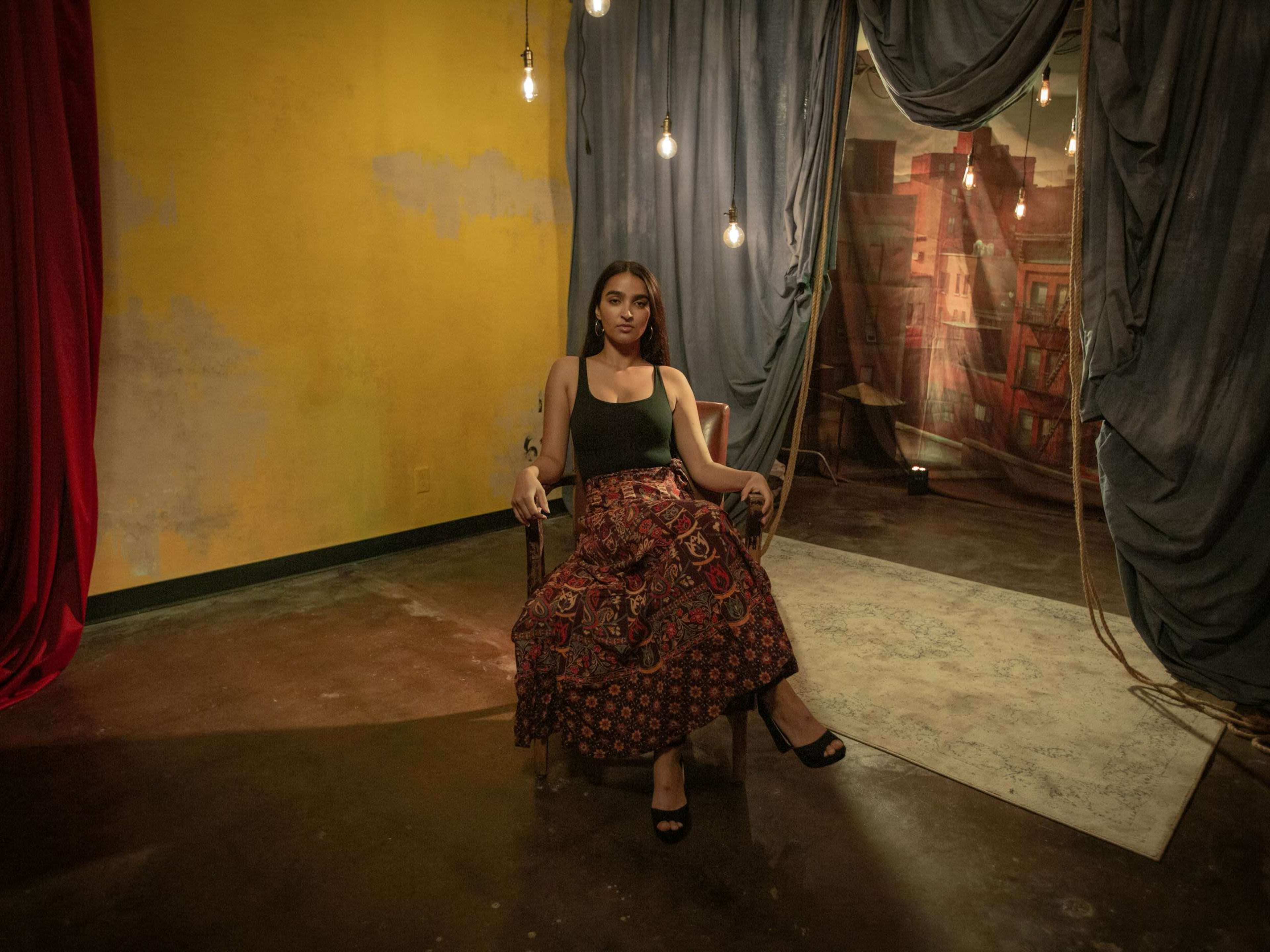 A person is sitting in a wooden chair in a dimly lit room with yellow and blue walls, draped fabric, and hanging light bulbs.