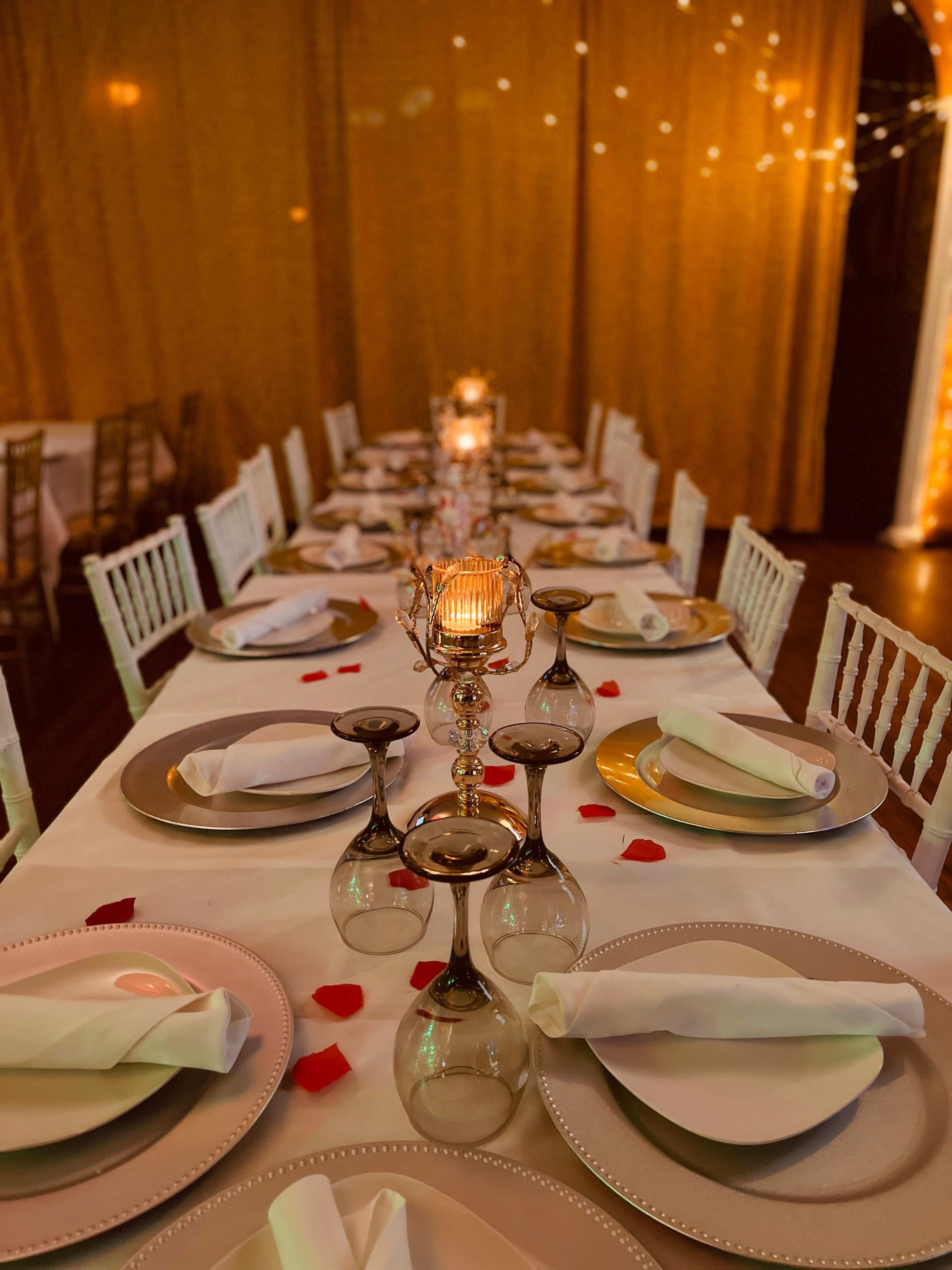 A long dining table is set with elegant plates, glassware, and candles, adorned with rose petals, under warm string lights and draped fabric.