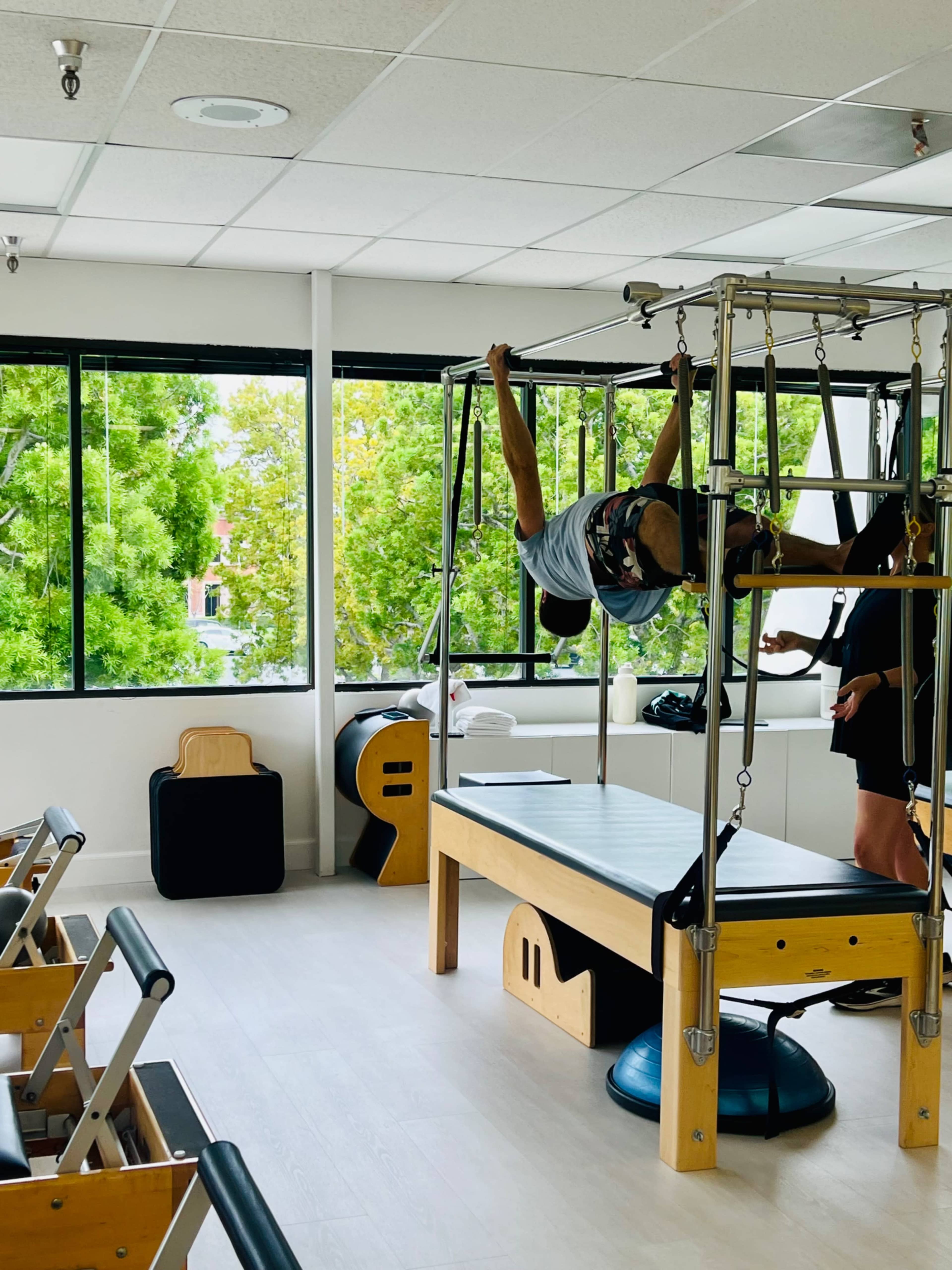 A person performs an exercise on a Pilates reformer in a well-lit studio with large windows overlooking greenery.