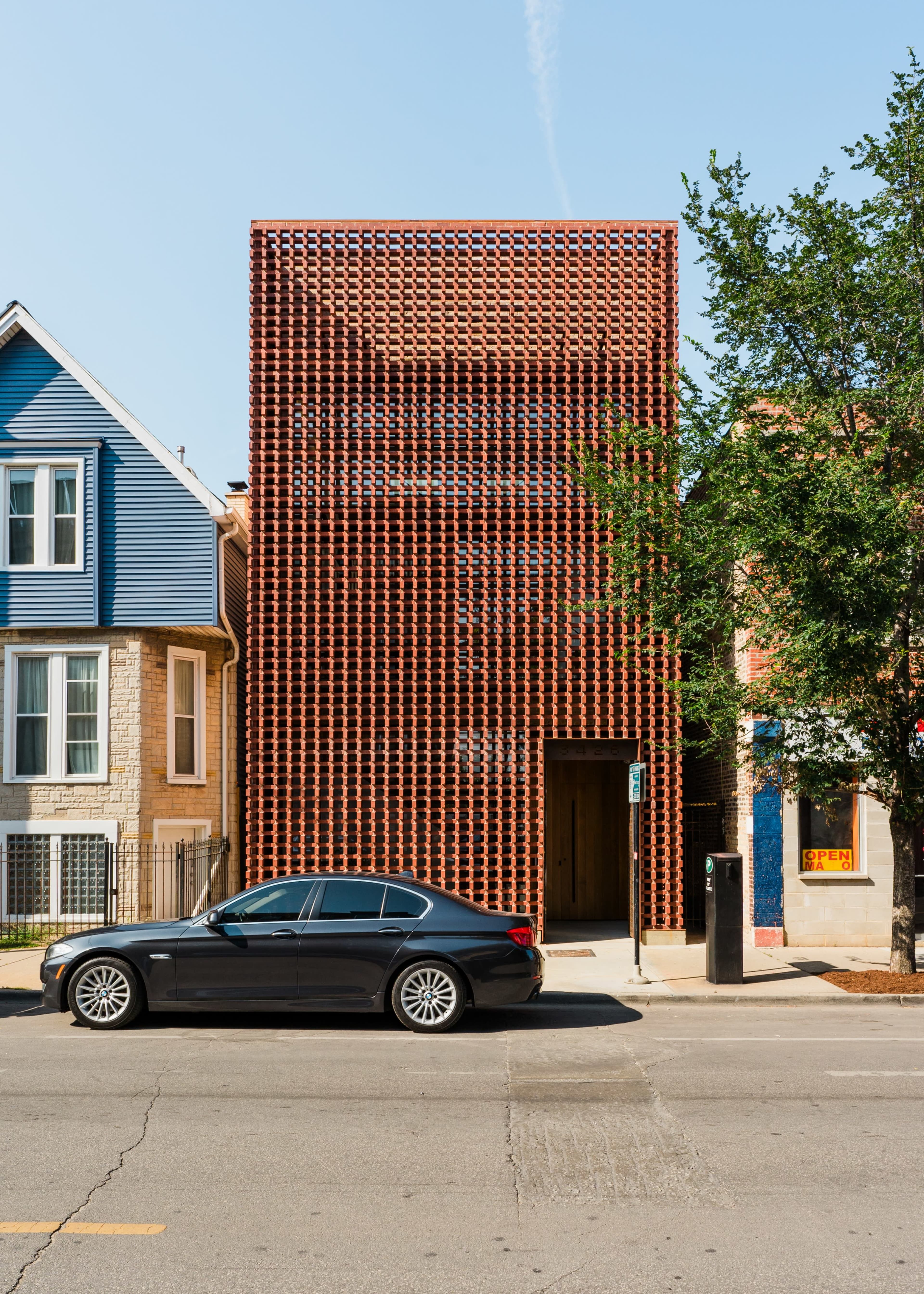 The image shows a modern building with a distinctive brick façade located on a city street, beside traditional residential houses and a parked black car.