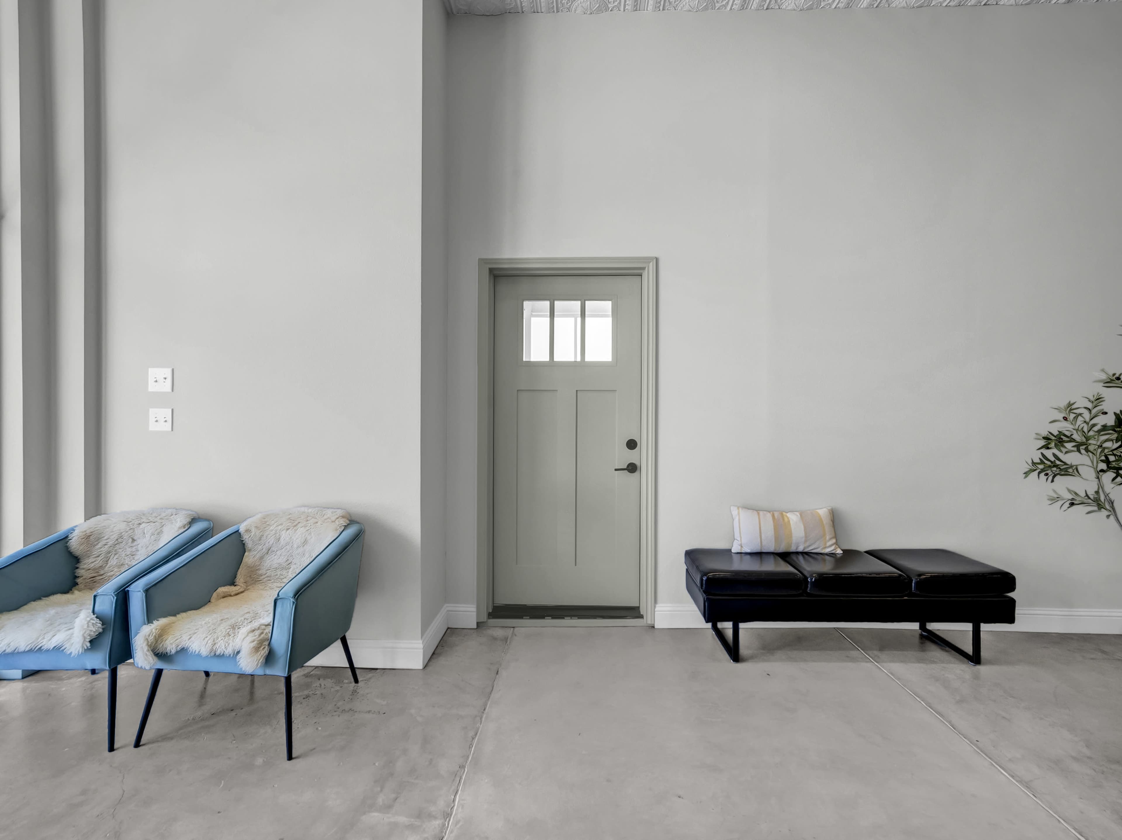 The image shows a minimalist interior space with two turquoise chairs and a black bench near a green door, set against a grey wall and concrete floor.
