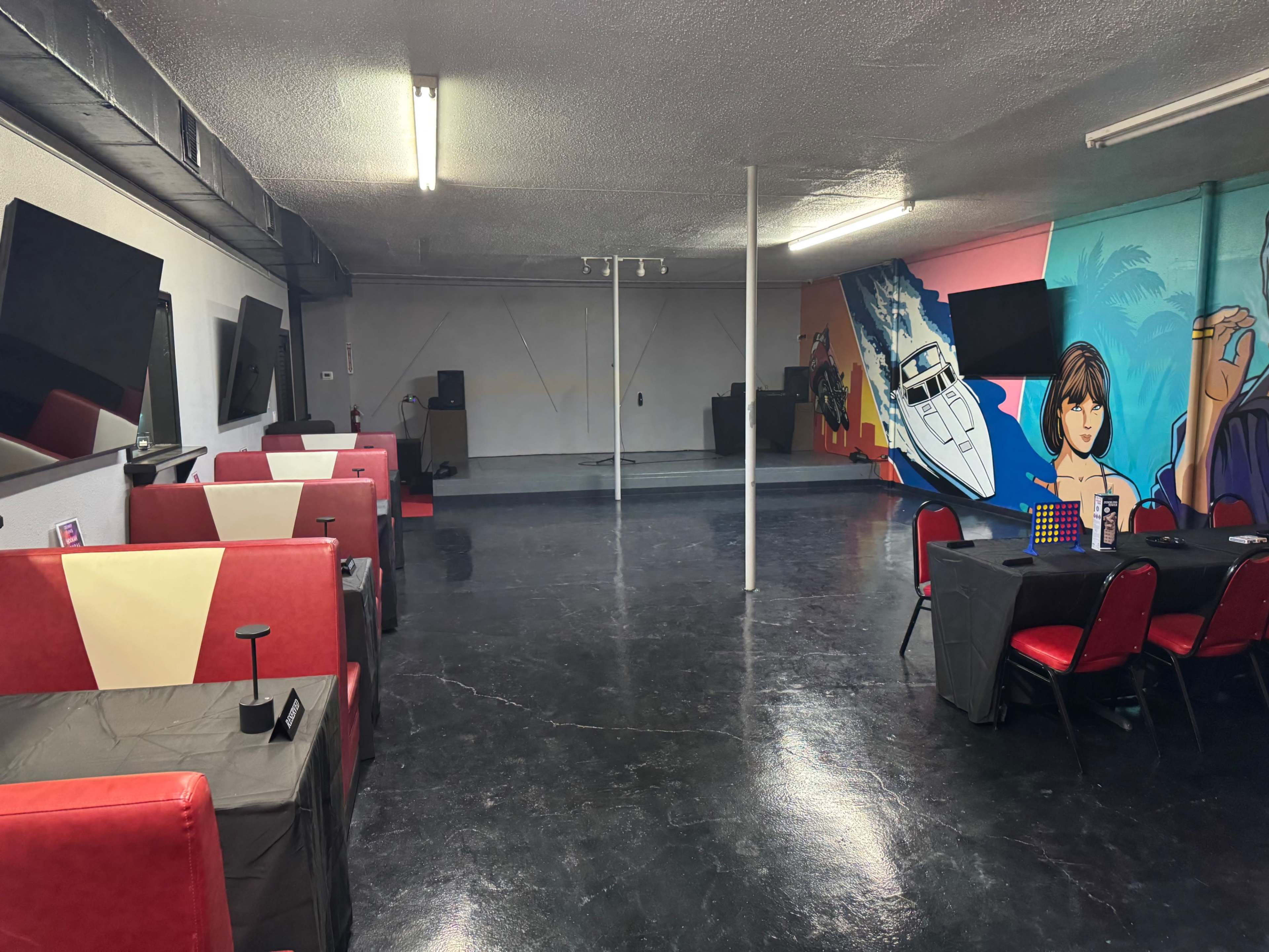 The image shows a spacious, brightly colored room with tables, chairs, and large TVs, featuring a mural on one wall.