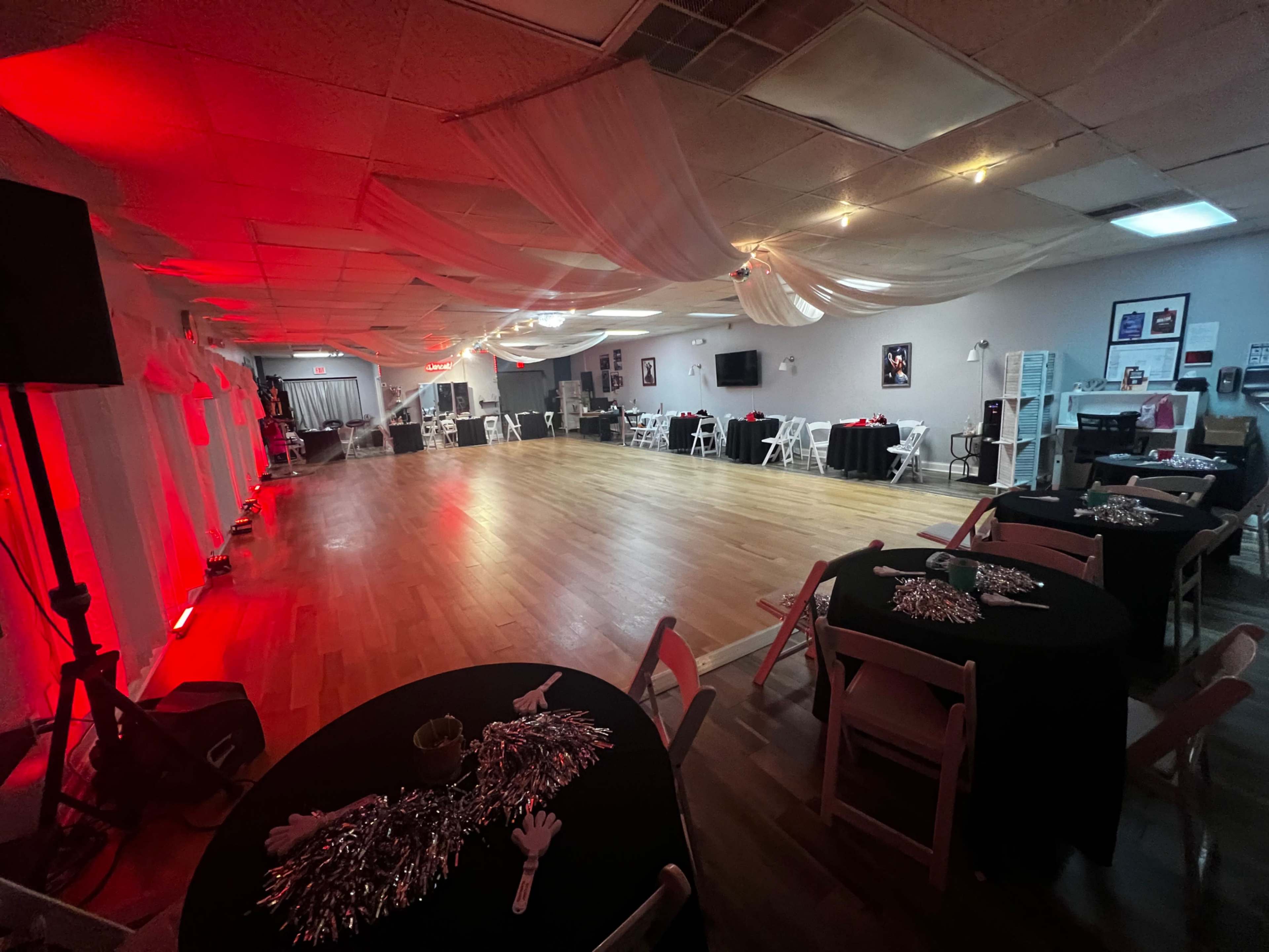 A spacious event hall with wooden flooring, black tables set with silver decorations, and soft red and white lighting.