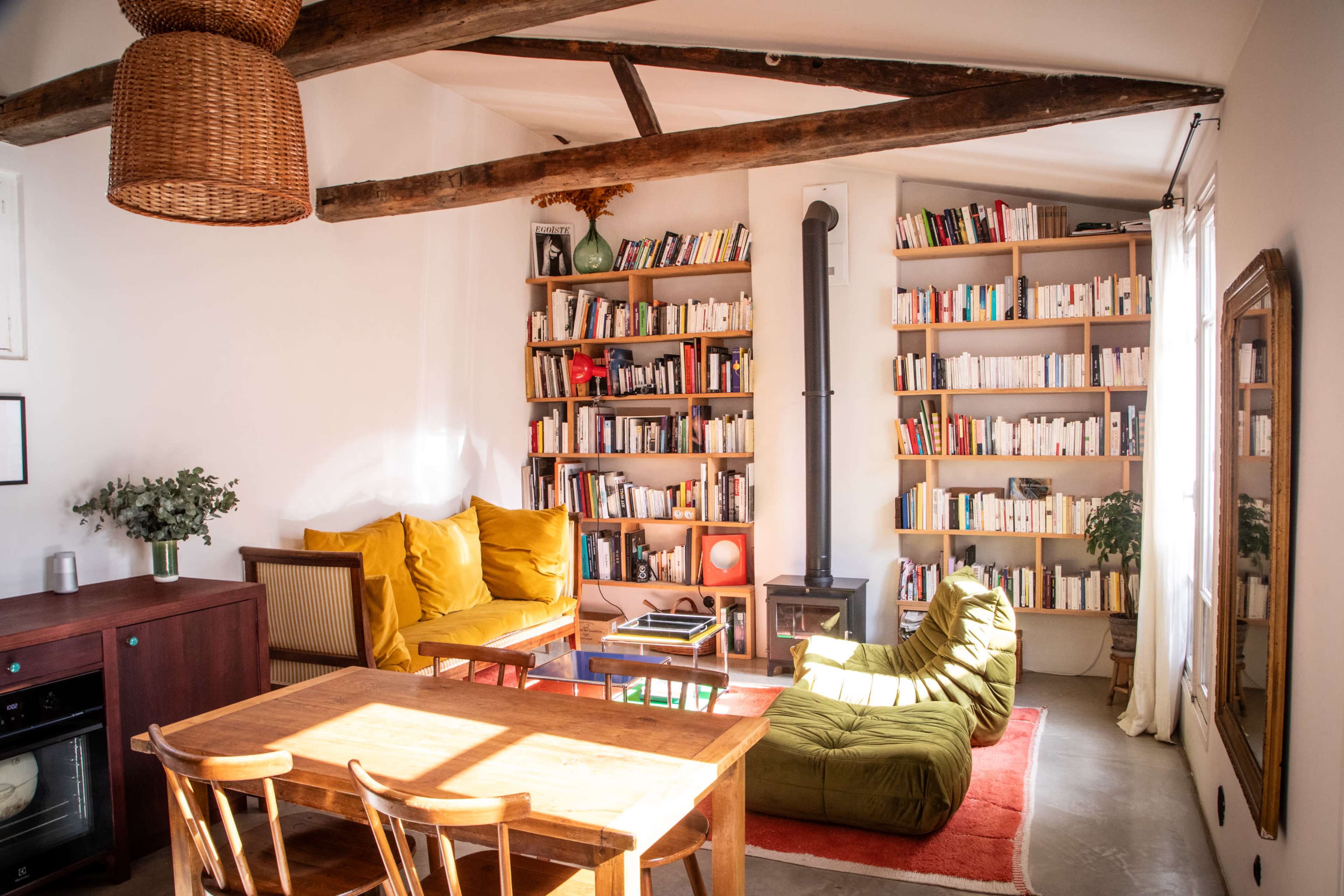 A cozy living room features a wooden table and chairs, a green beanbag chair, shelves filled with books, and sunlight streaming through a window.