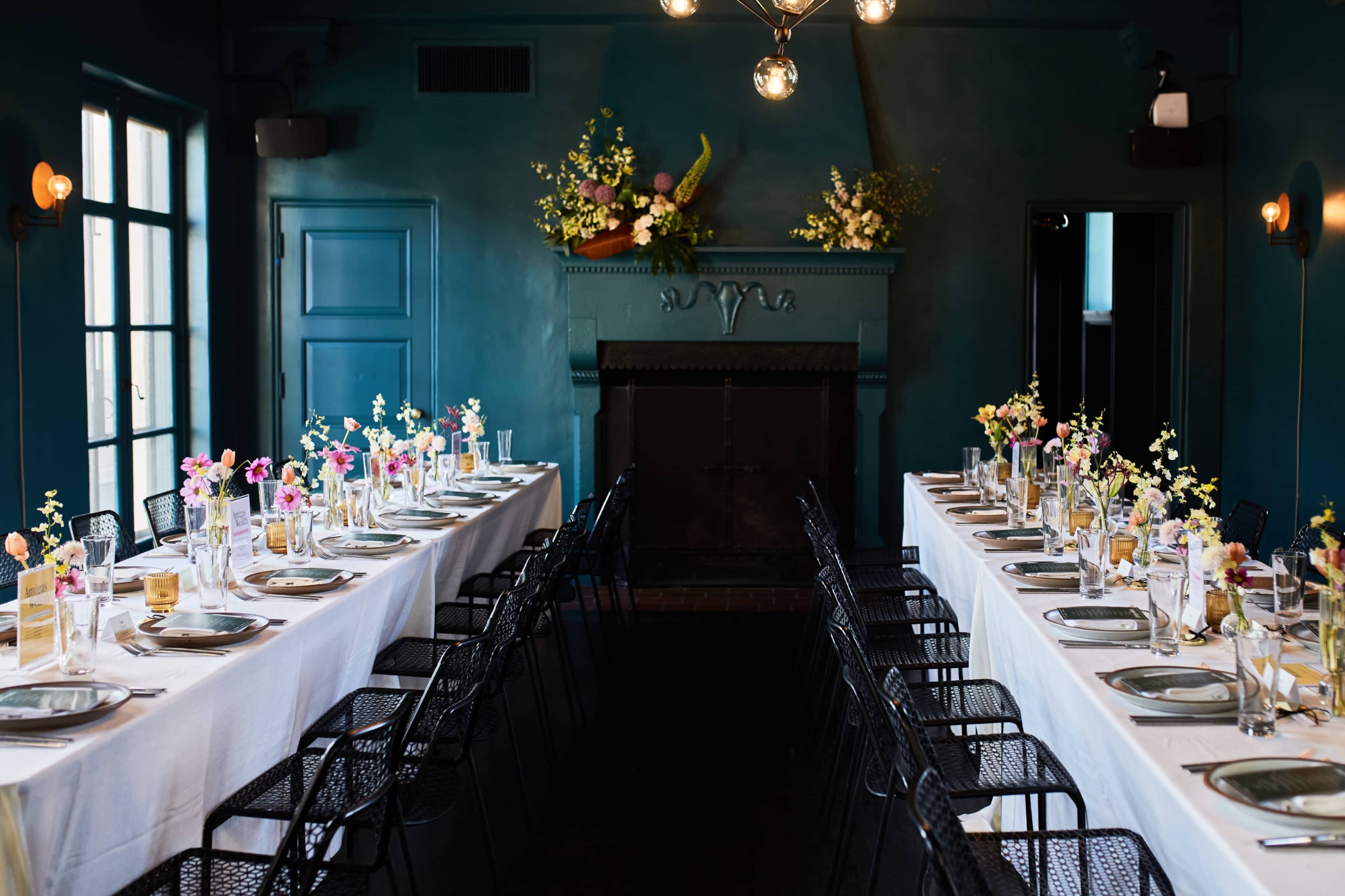 A long banquet table is set with elegant place settings and floral centerpieces in a dimly lit room with blue walls and a decorative fireplace.