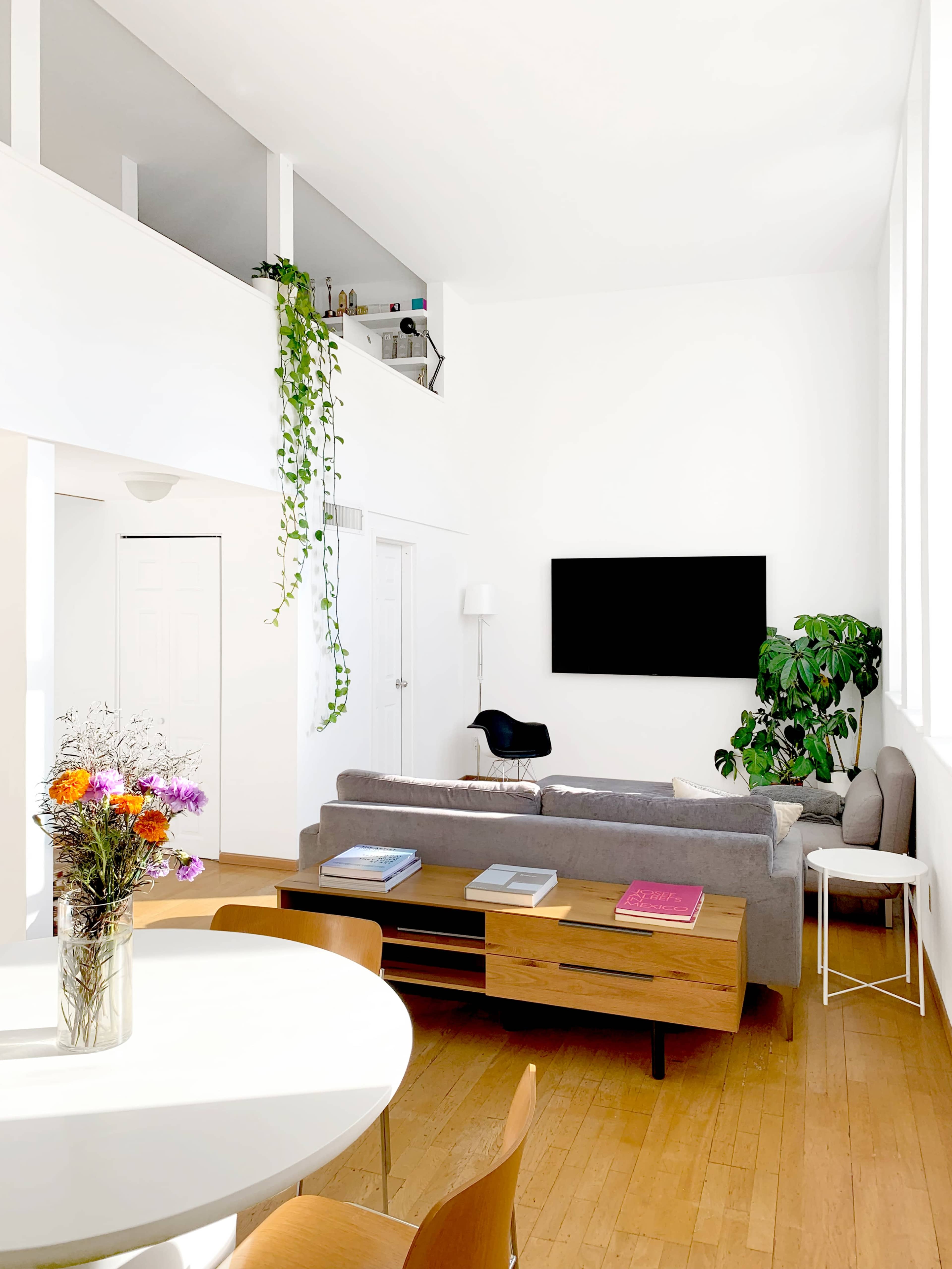 The image depicts a bright living room featuring a gray sofa, a wooden coffee table, a wall-mounted TV, and large windows that allow natural light to enter the space.