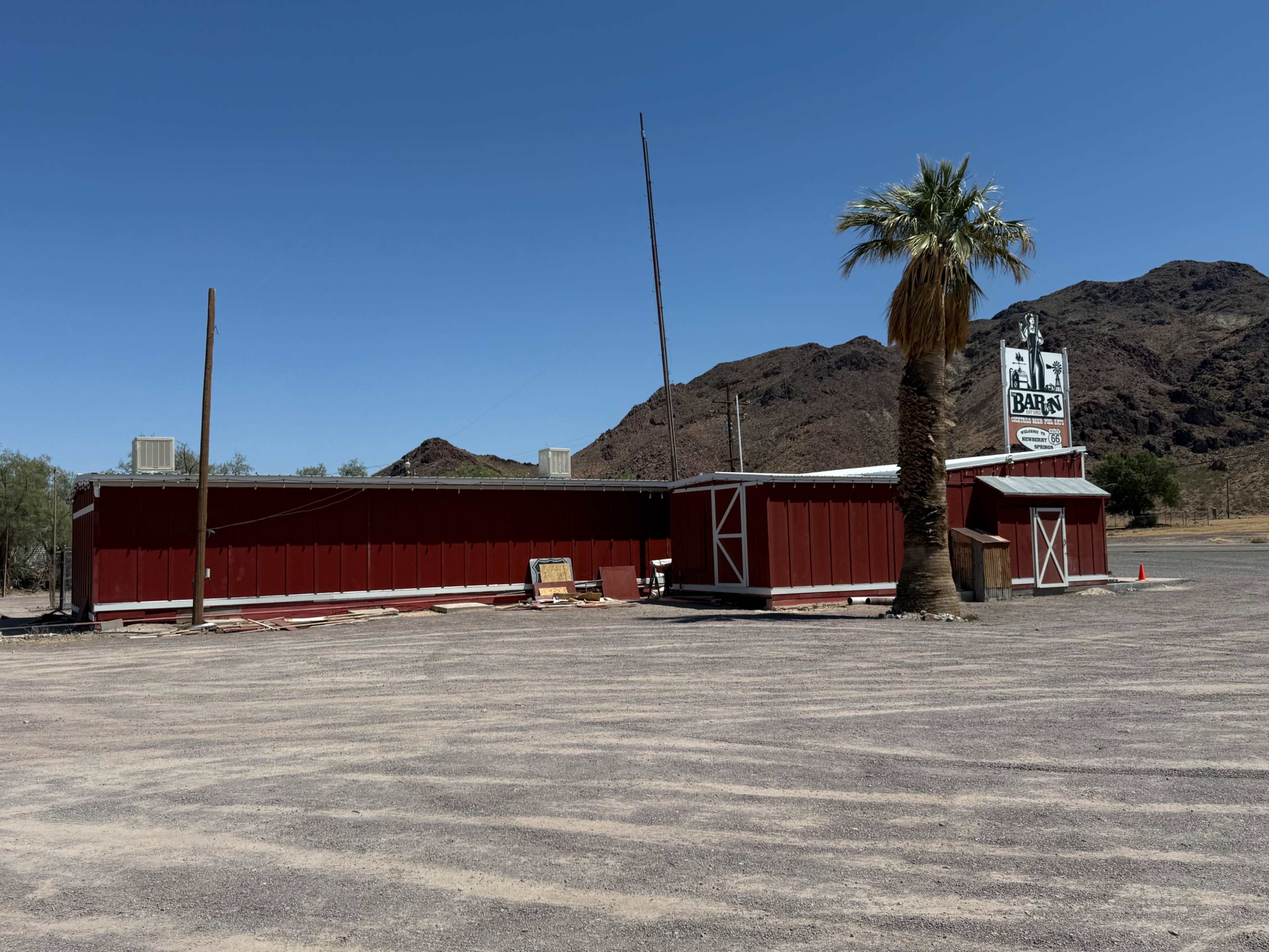 A red barn-style building stands beside a palm tree under a clear blue sky, with mountains in the background.