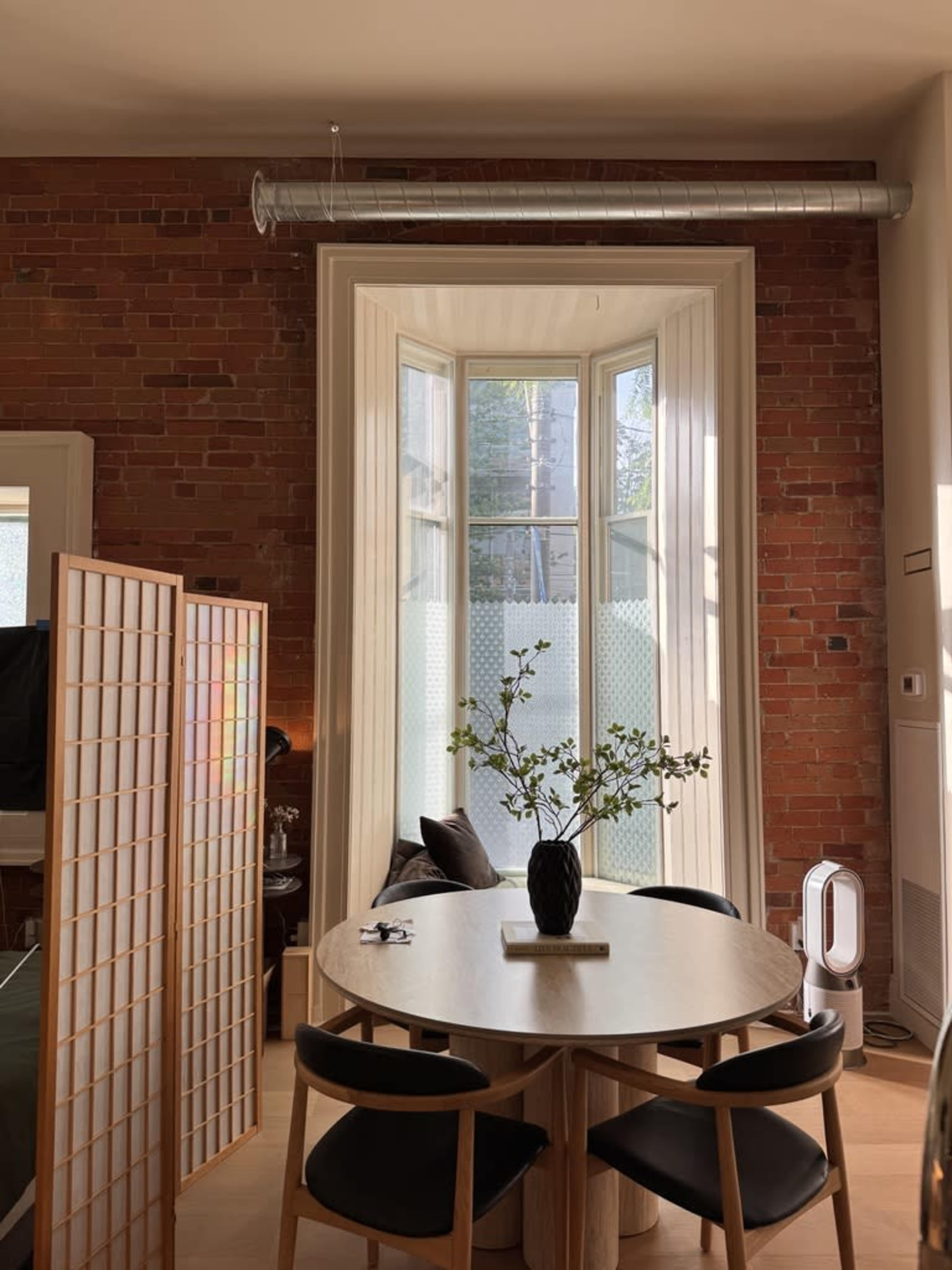 The image shows a dining area with a round wooden table and black chairs, positioned next to a large bay window with brick walls and a plant vase in the center.