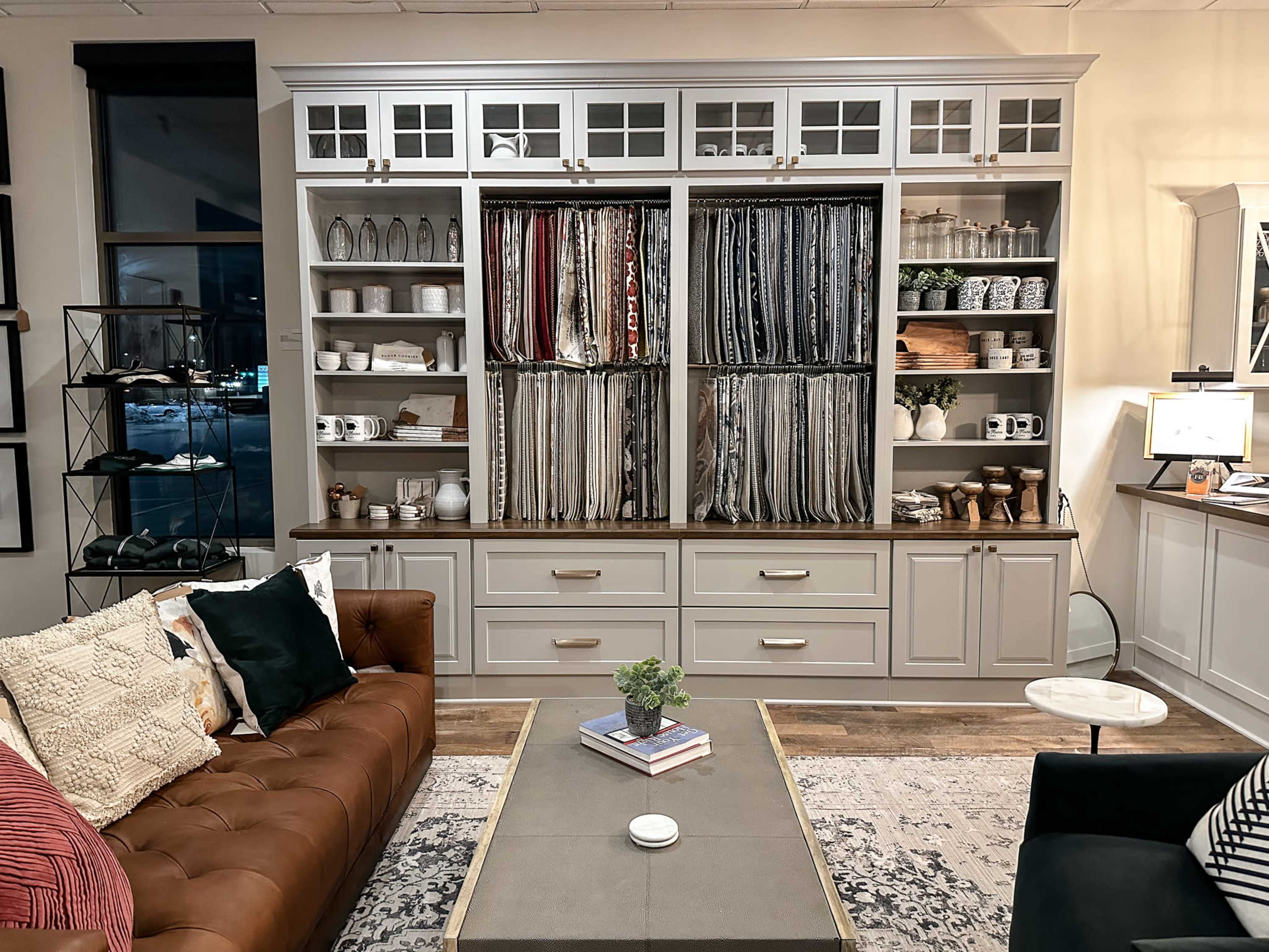 A cozy living room setting featuring a brown leather sofa, a central coffee table, and a storage unit filled with various fabric samples, dishes, and decorative items.