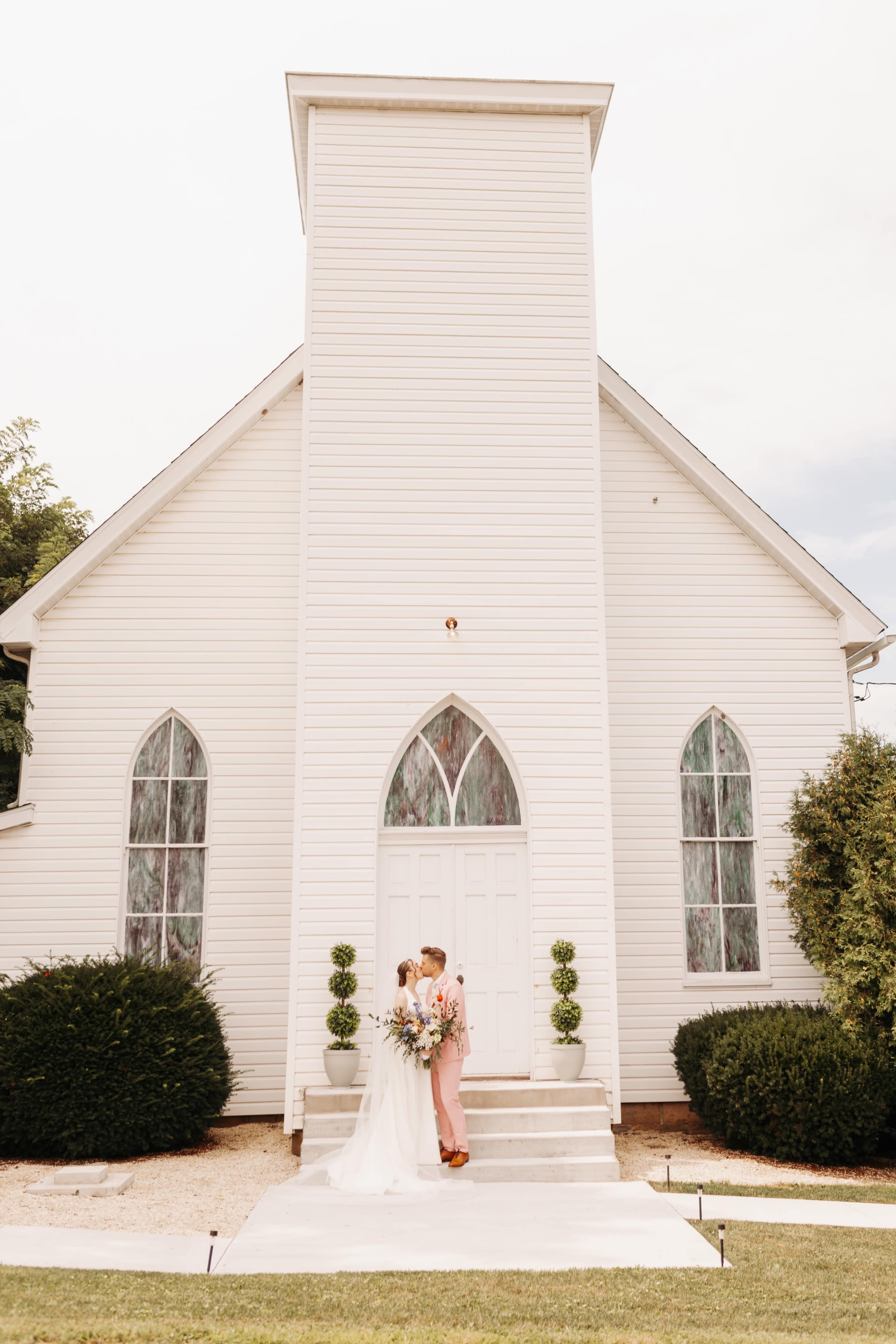 A couple stands in front of a white church with large arched windows, holding a bouquet and dressed in formal attire.