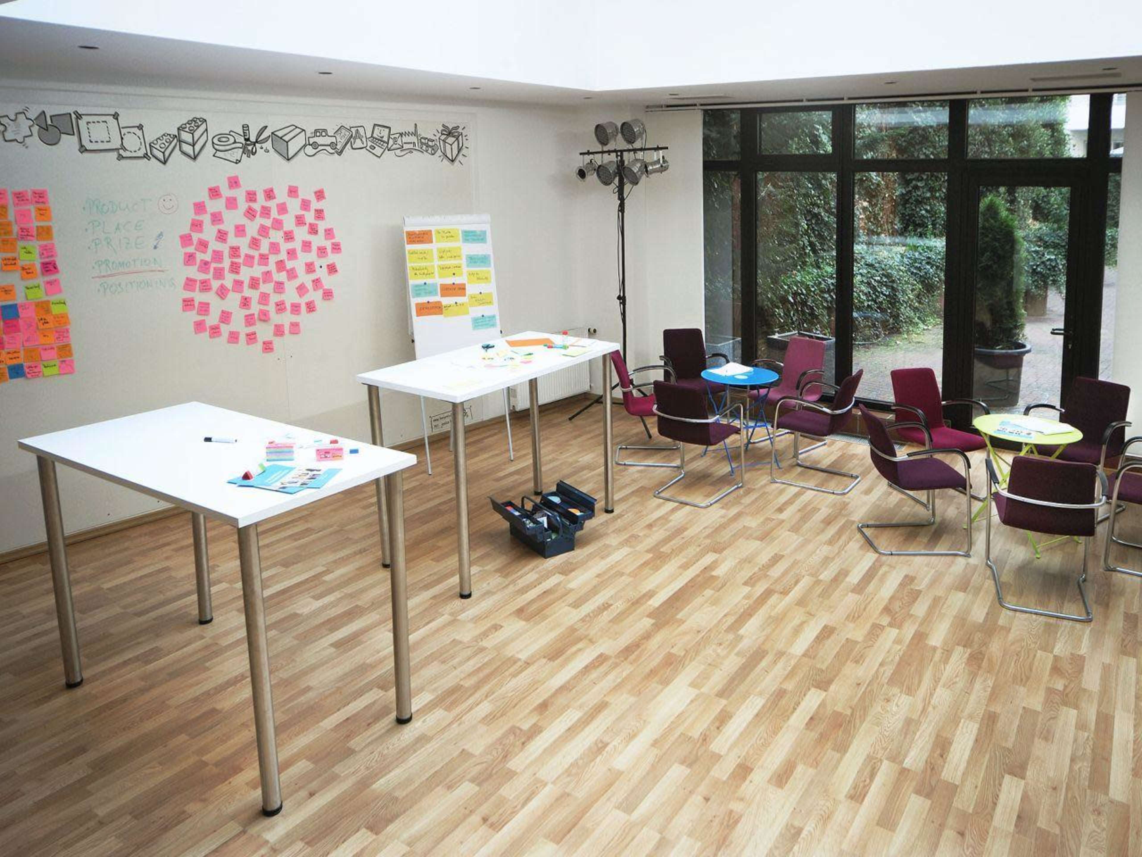 A spacious meeting room features several white tables, colorful sticky notes on the walls, and a cluster of chairs arranged around small tables.