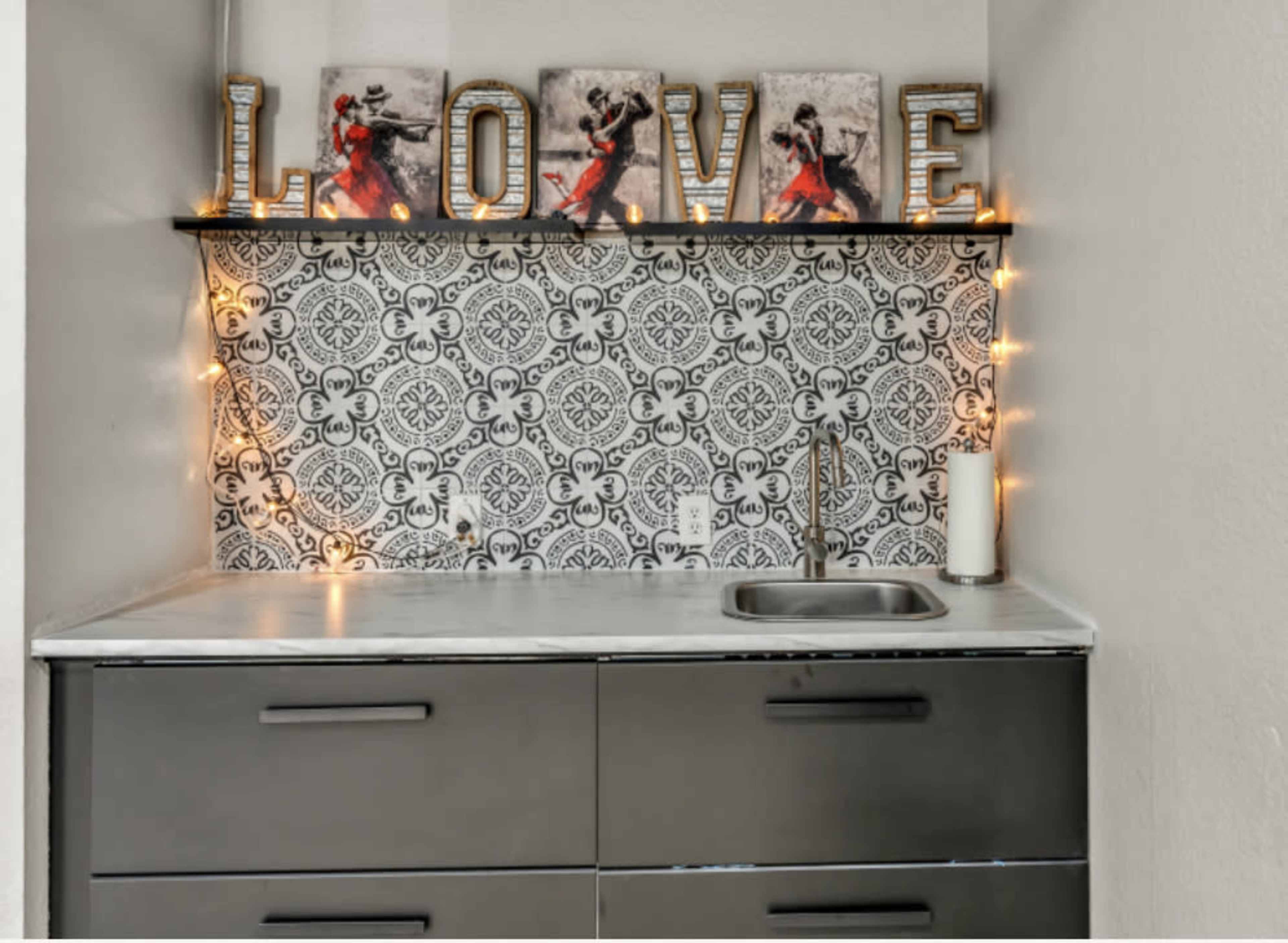 A small kitchen area with a sink, dark cabinets, decorative letters spelling "LOVE," and a patterned backsplash illuminated by string lights.
