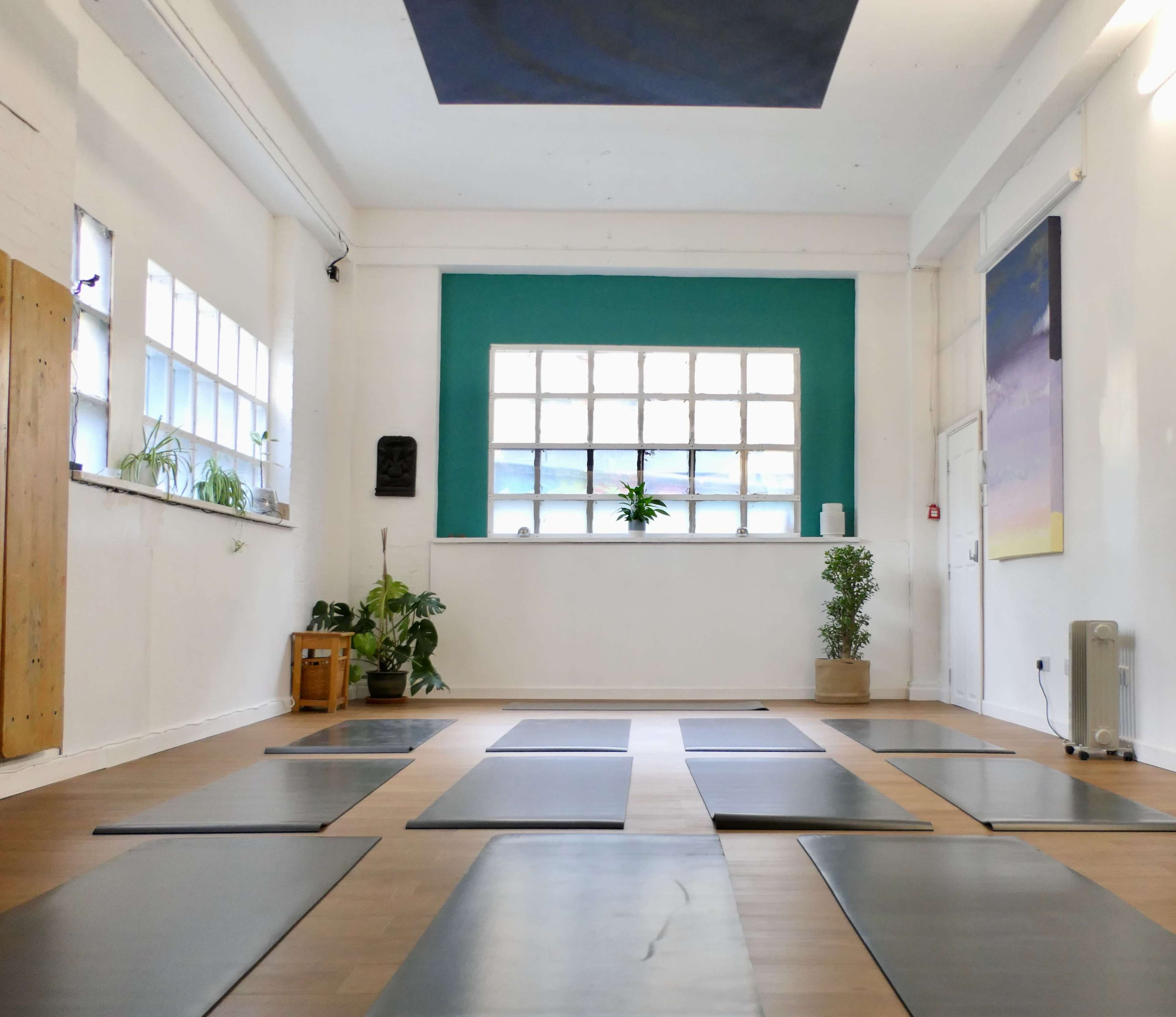 A spacious yoga studio with several yoga mats arranged on a wooden floor, large windows, and green wall accents.