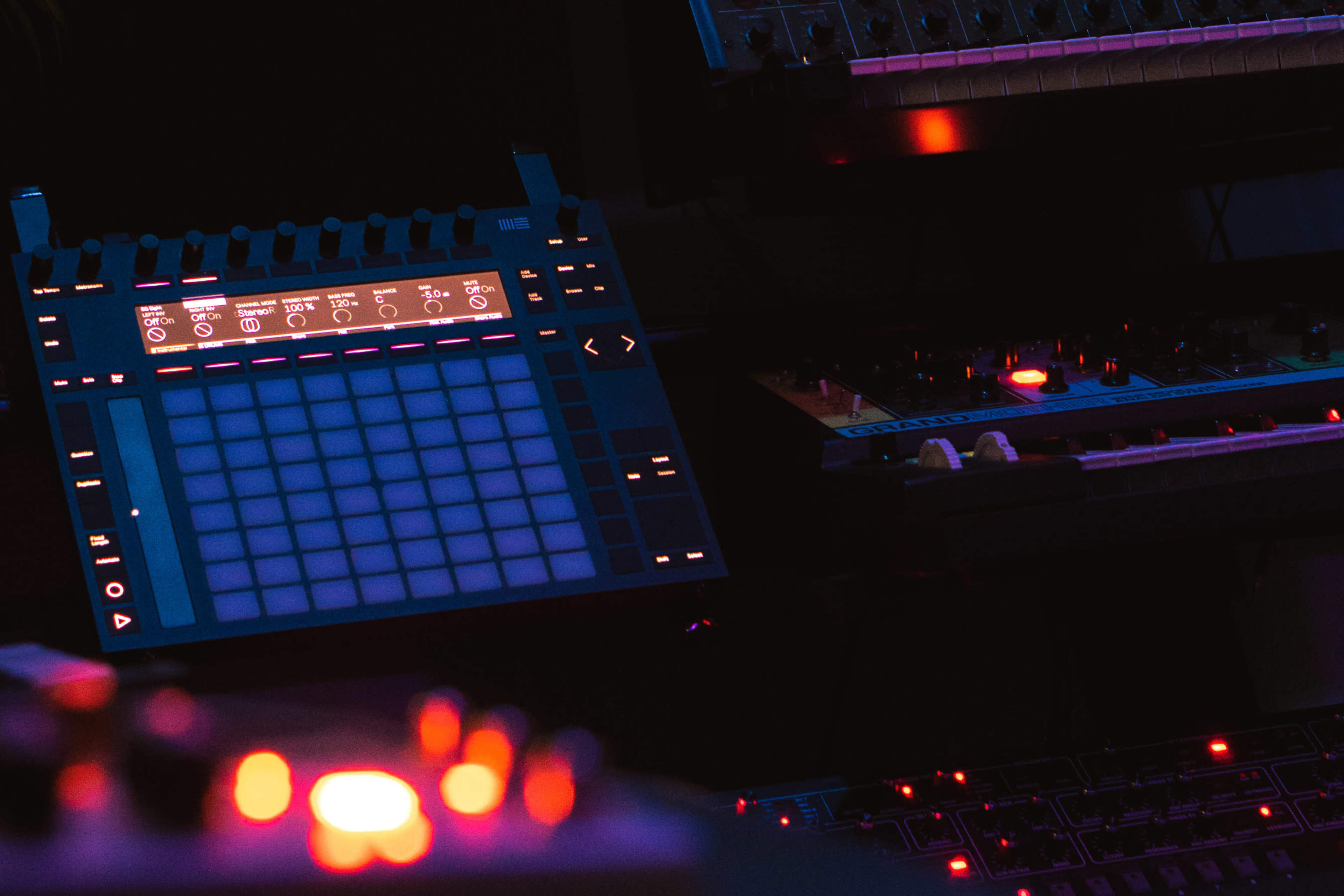 The image shows a close-up of electronic music equipment, featuring a grid pad controller and various synthesizers with illuminated buttons and controls.