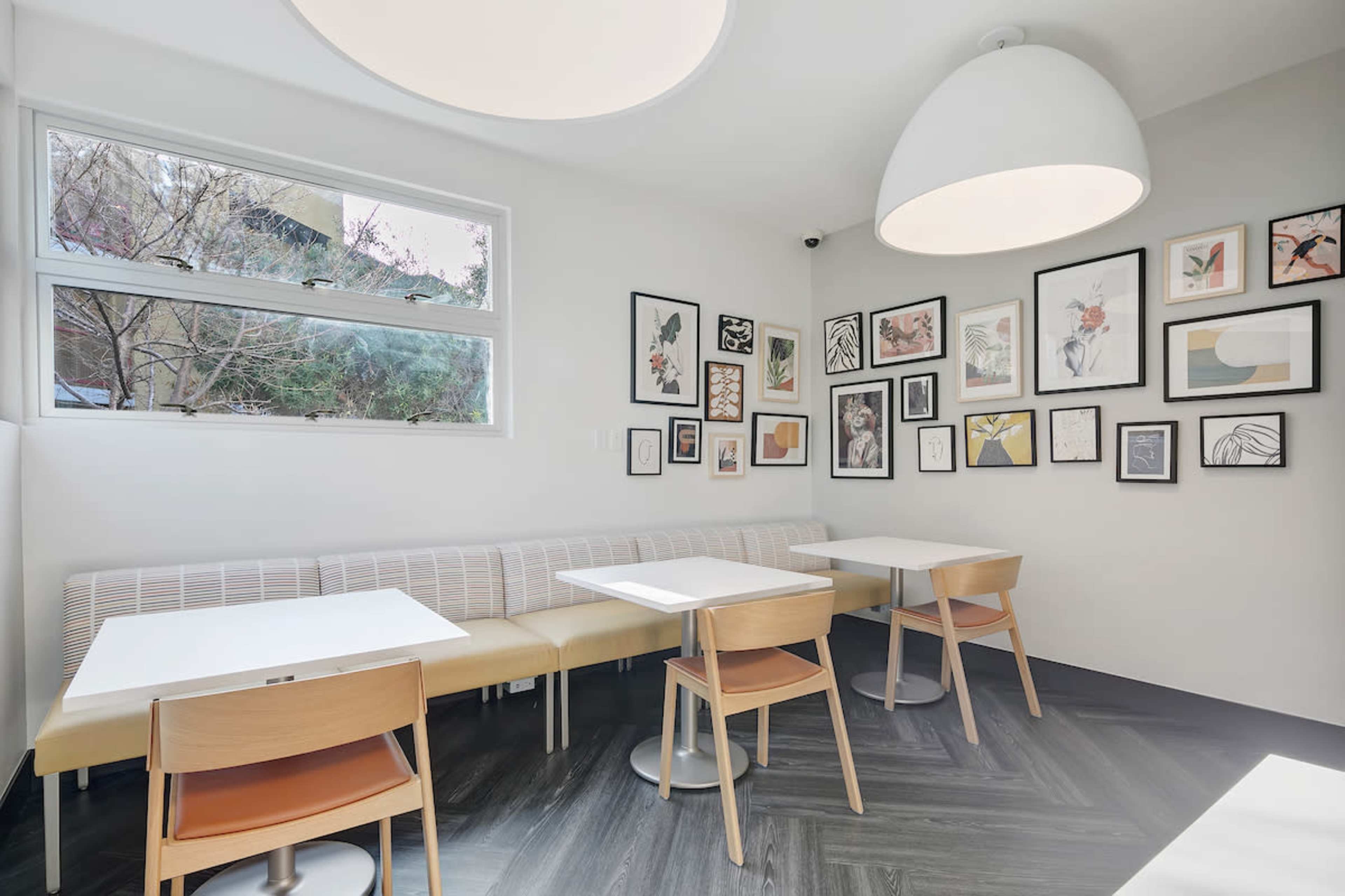 A small café interior with a minimalist design featuring a bench along the wall and tables with chairs arranged in a corner, surrounded by framed artwork on the walls.