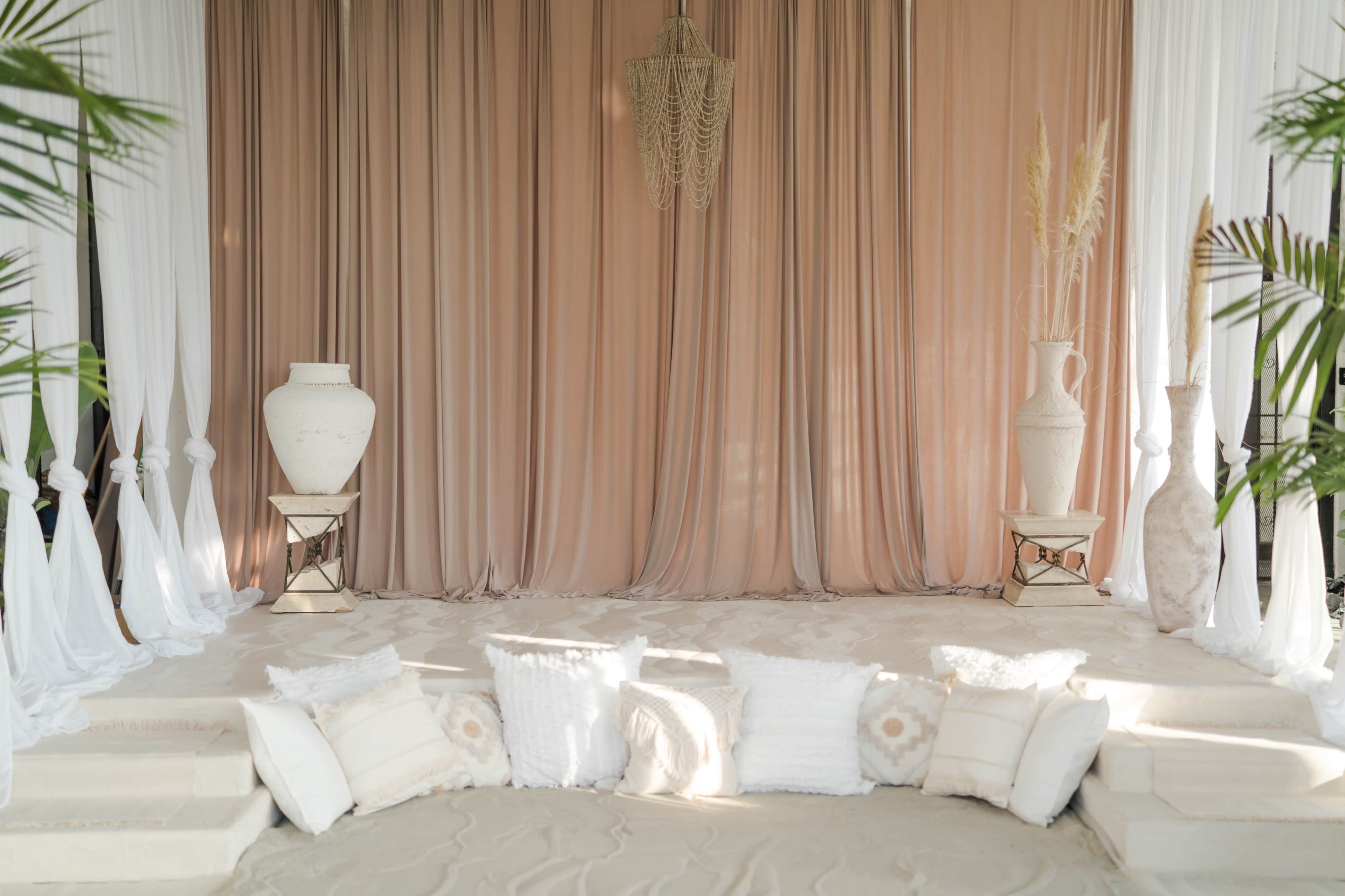 A stage is set with draped fabric in beige tones, flanked by large vases and cushions arranged on a sandy floor.