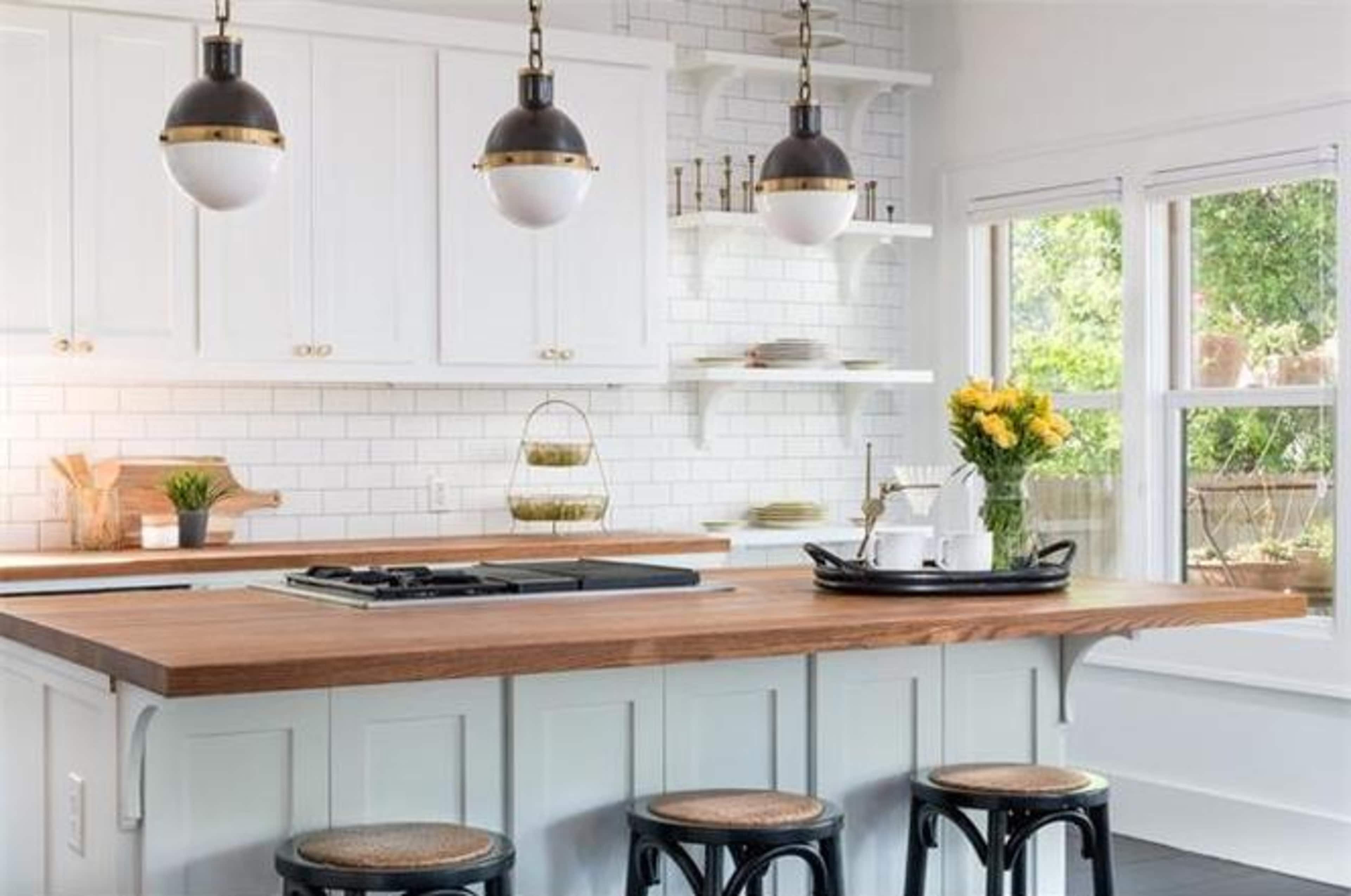 The kitchen features a large wooden island with a stovetop, white cabinetry, and three black stools, complemented by pendant lights and a window overlooking greenery.