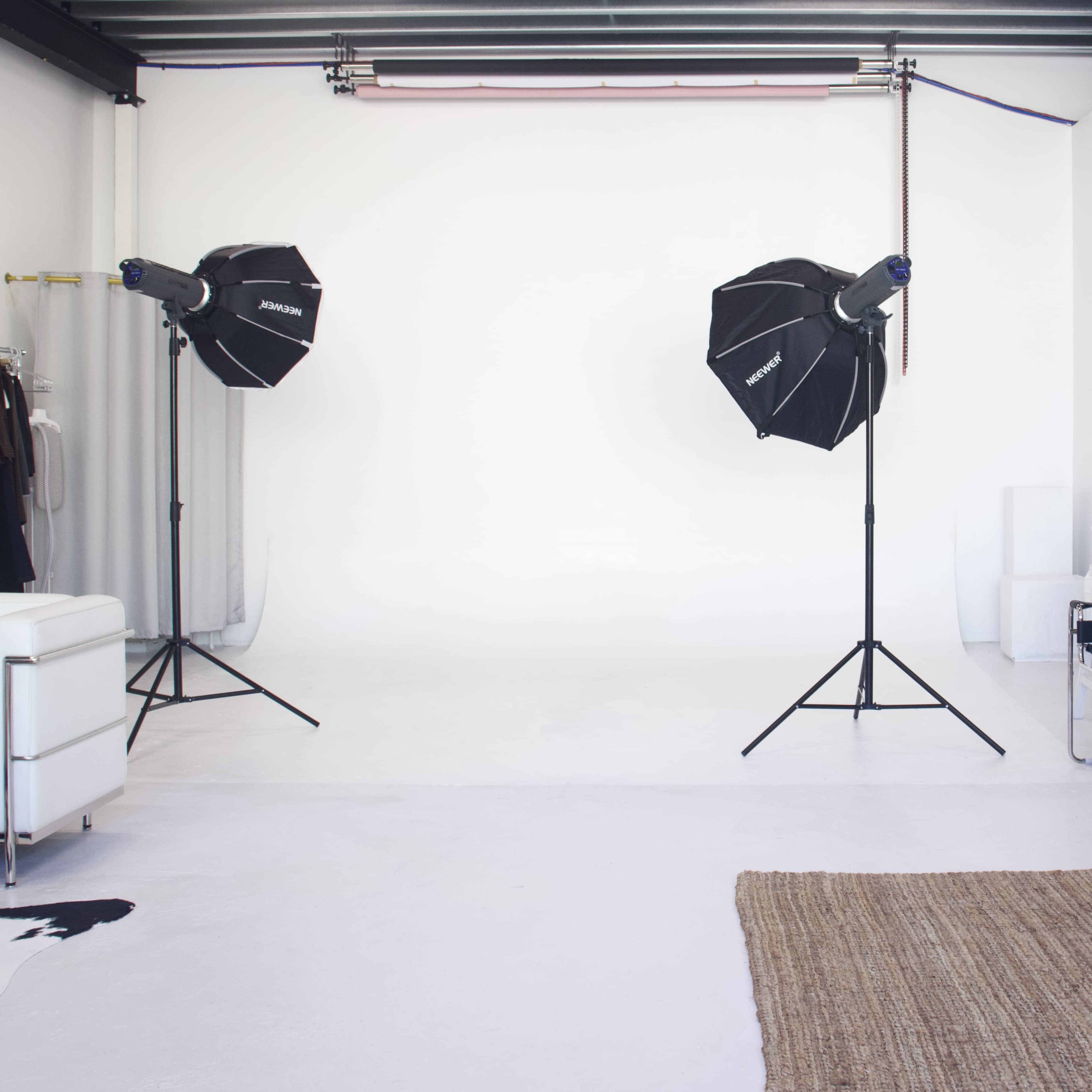The image shows a minimalistic photo studio featuring two lighting setups and a white backdrop on a clean, light-colored floor.