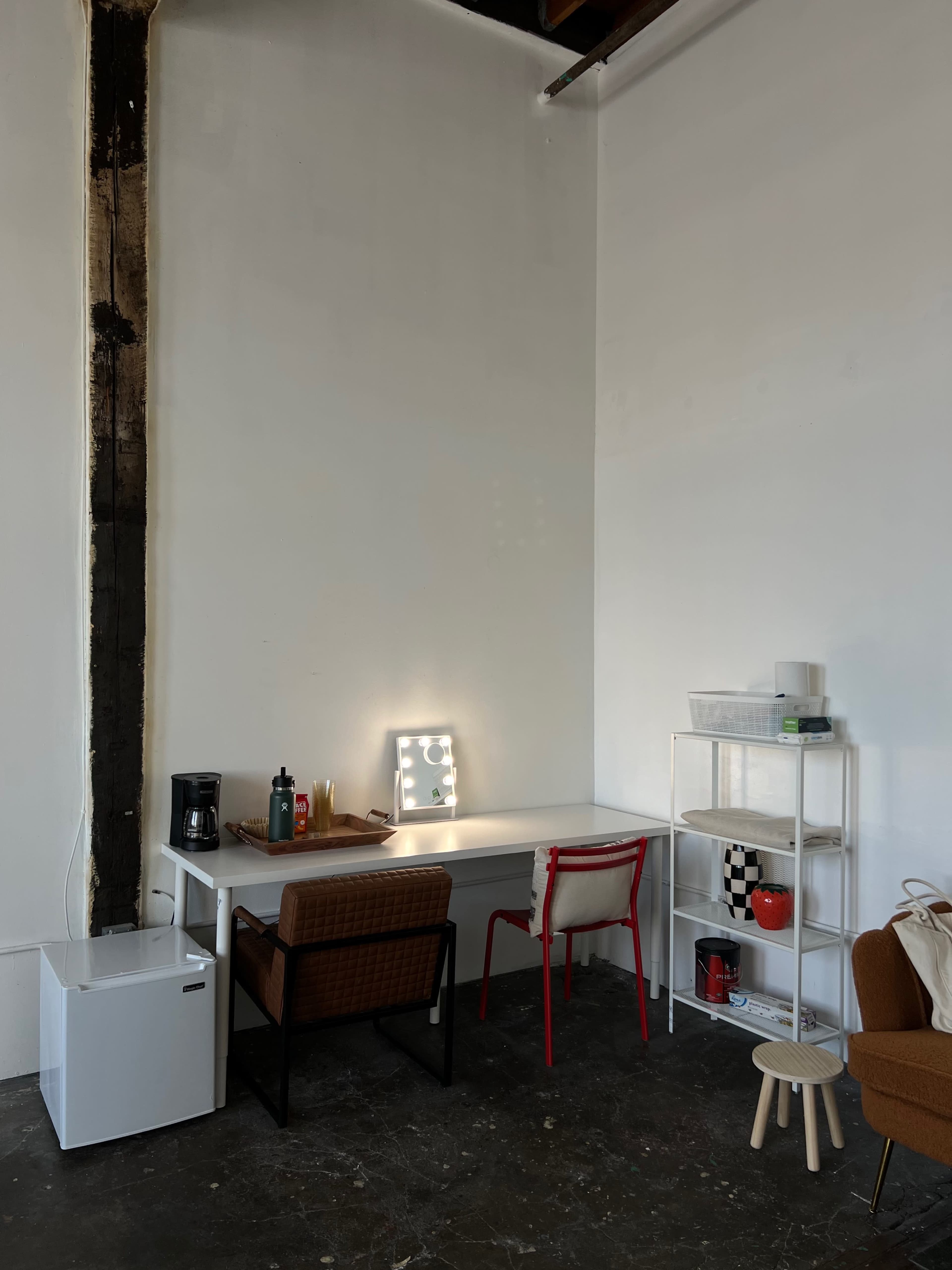 A minimal workspace features a white table with two chairs, a coffee maker, and shelves, set against a stark white wall.