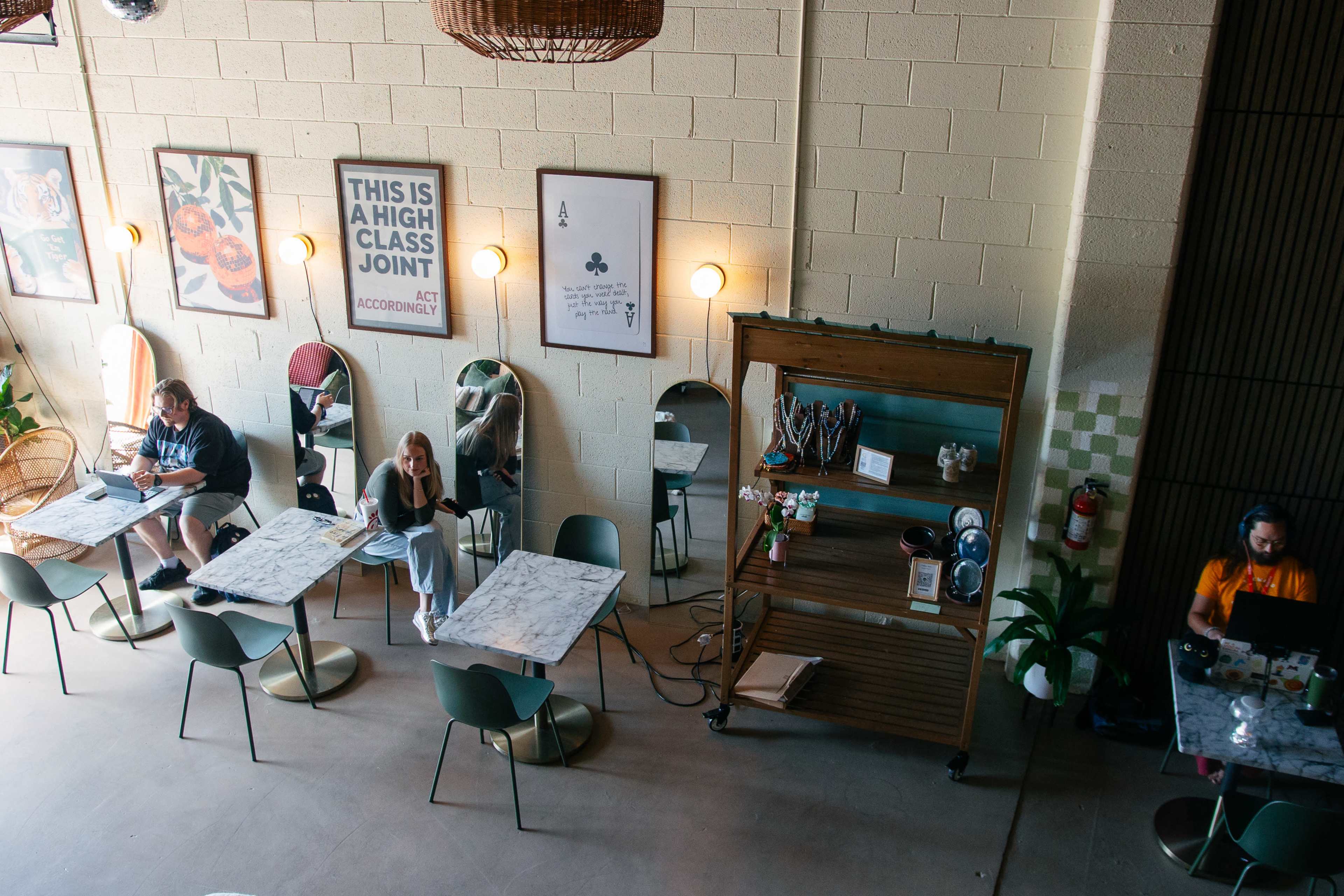 The image shows a cozy cafe interior with several patrons seated at tables, decorative mirrors on the walls, and a wooden shelving unit displaying coffee-making equipment.