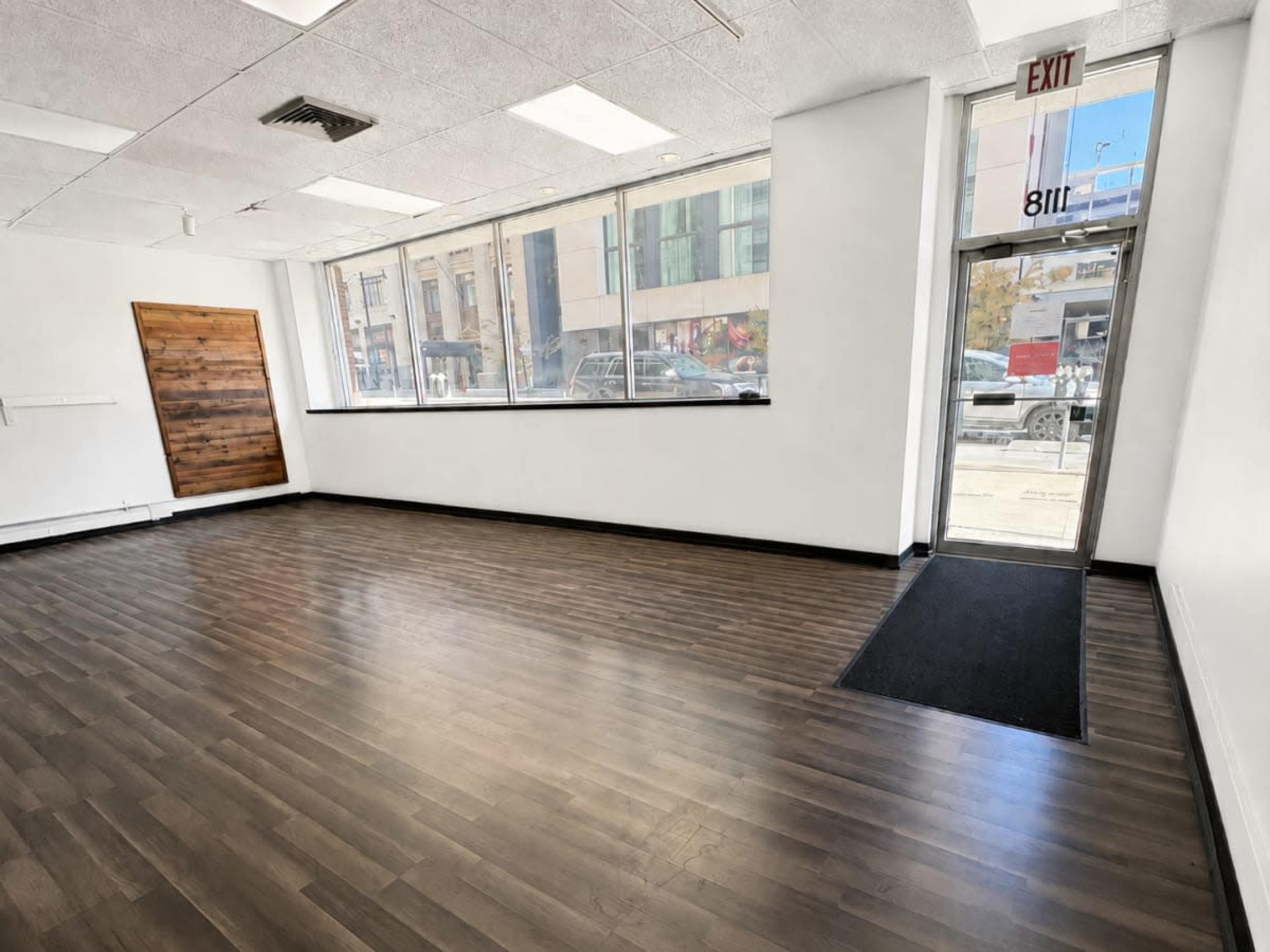 The image shows an empty retail space with wooden flooring, large front windows, and a door leading outside.