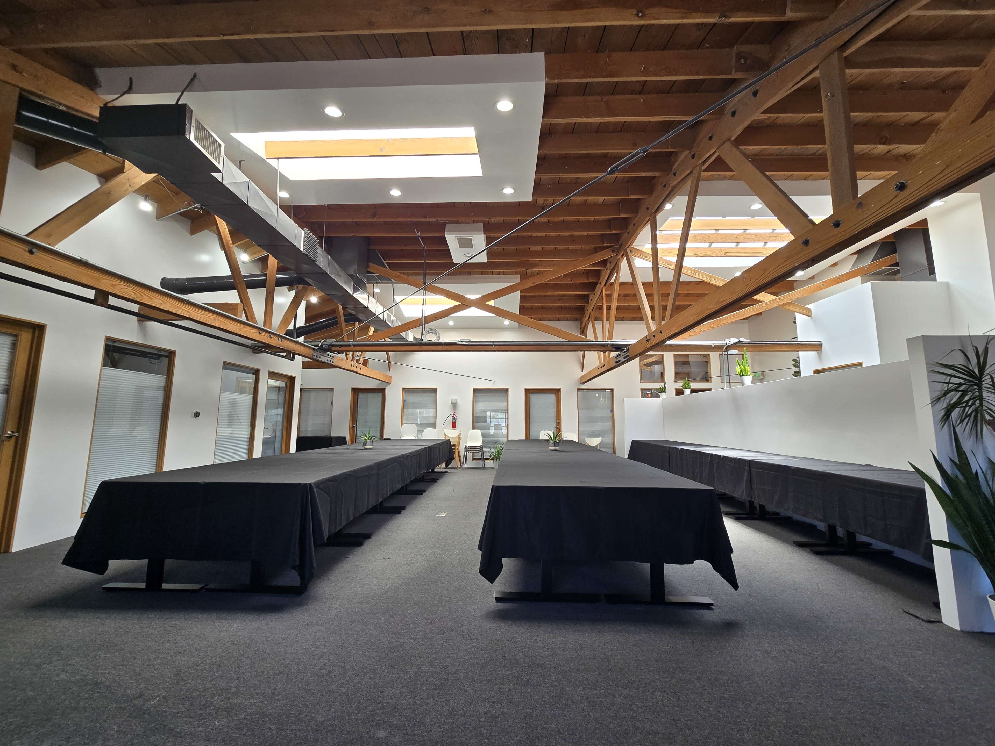 The image shows a spacious, modern meeting room with several long tables covered in black tablecloths and exposed wooden beams overhead.